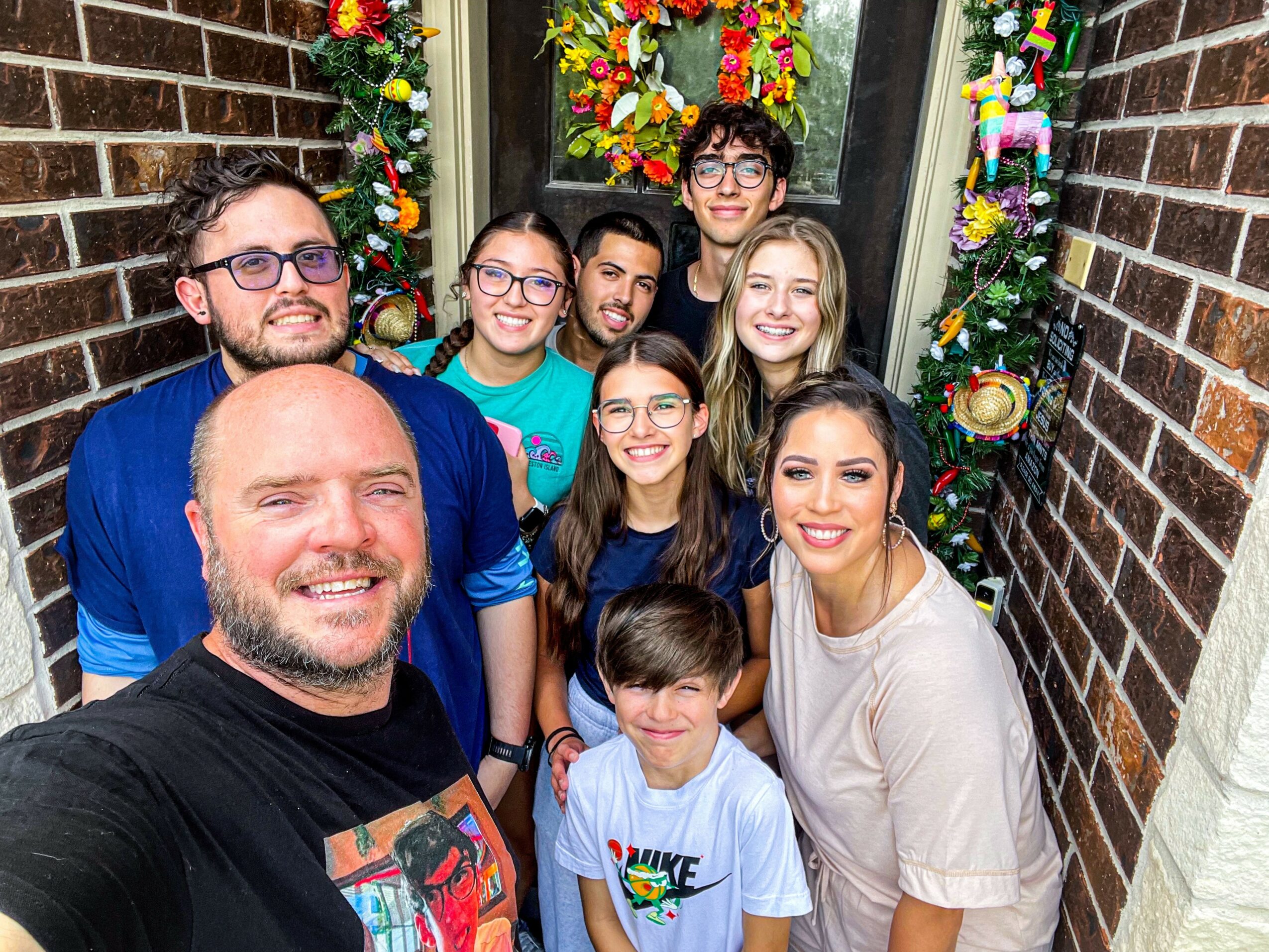 Group of nine people, including children and adults, smiling in front of decorated window with Christmas ornaments, brick walls on sides.