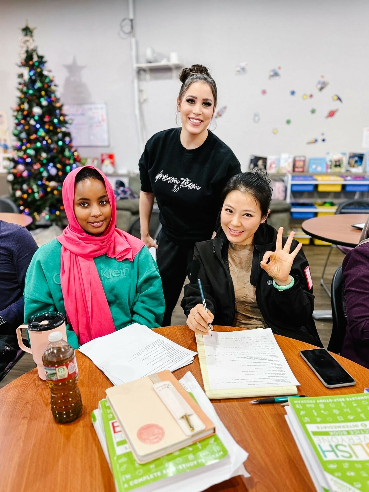 Three women sitting at a table in a decorated room with a Christmas tree, smiling and making peace signs.