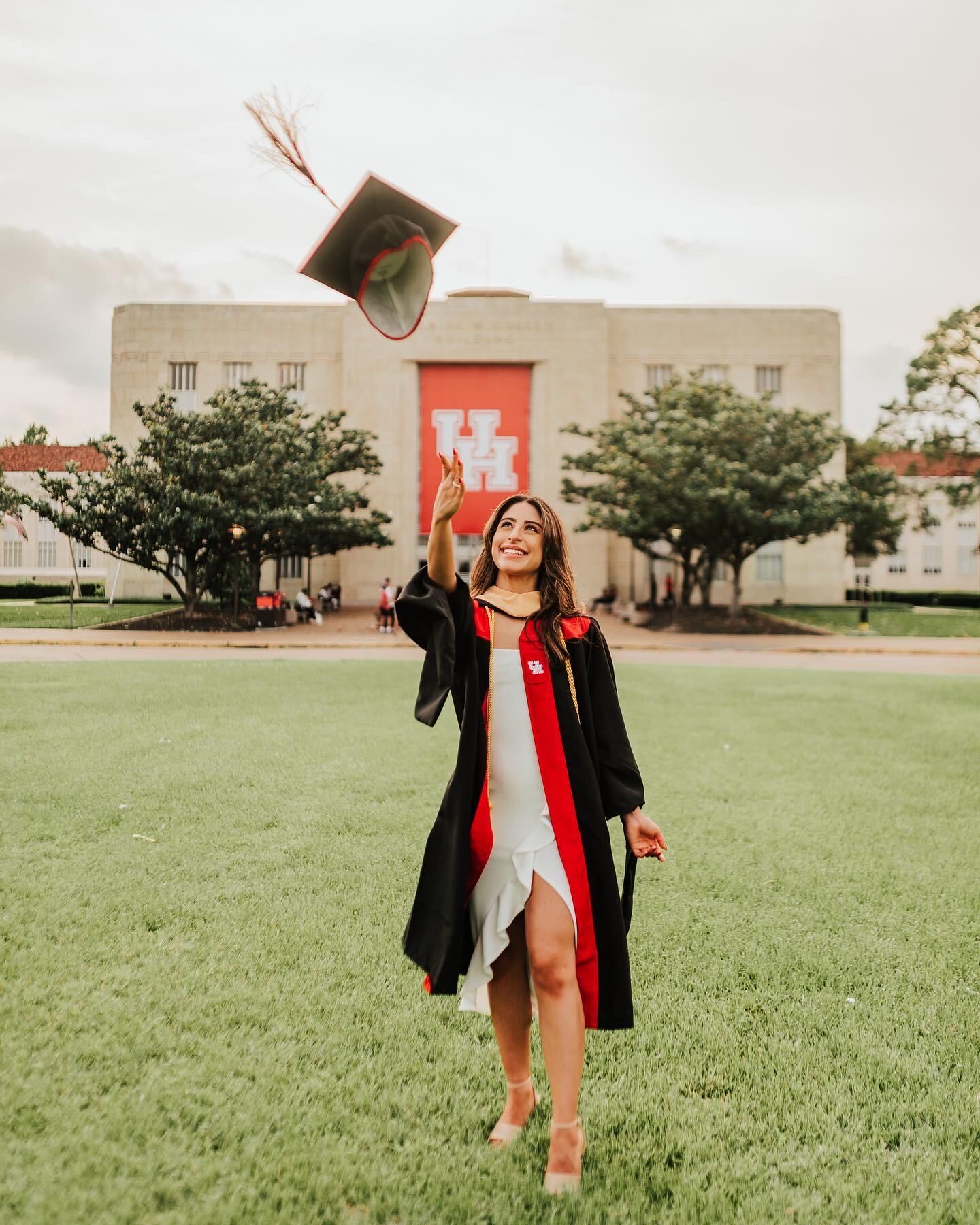 Graduate in cap and gown tossing cap in the air on campus lawn with building and trees in background.