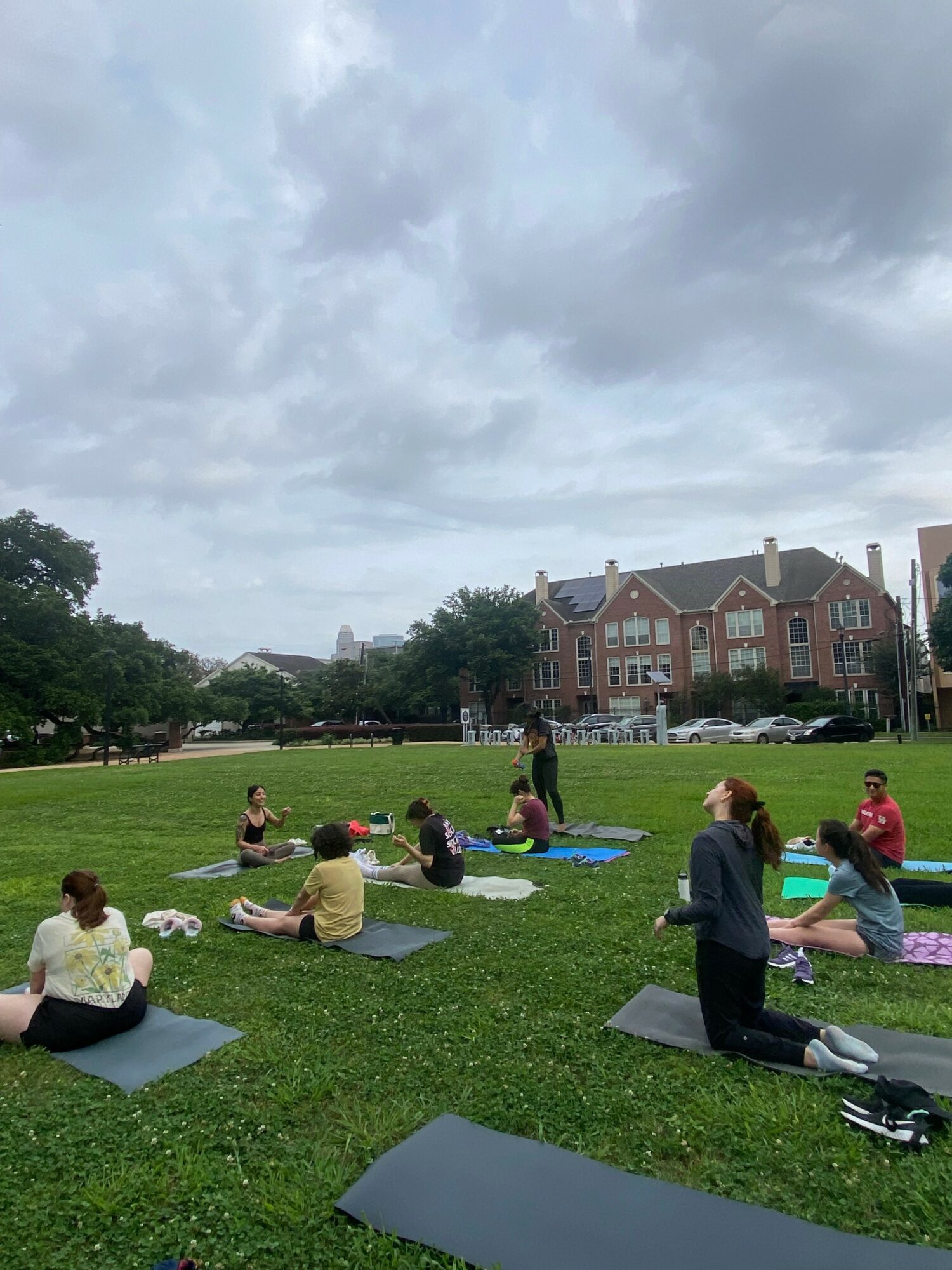 People practicing yoga outdoors on mats in a park with green grass and trees, under a cloudy sky, with buildings in the background.