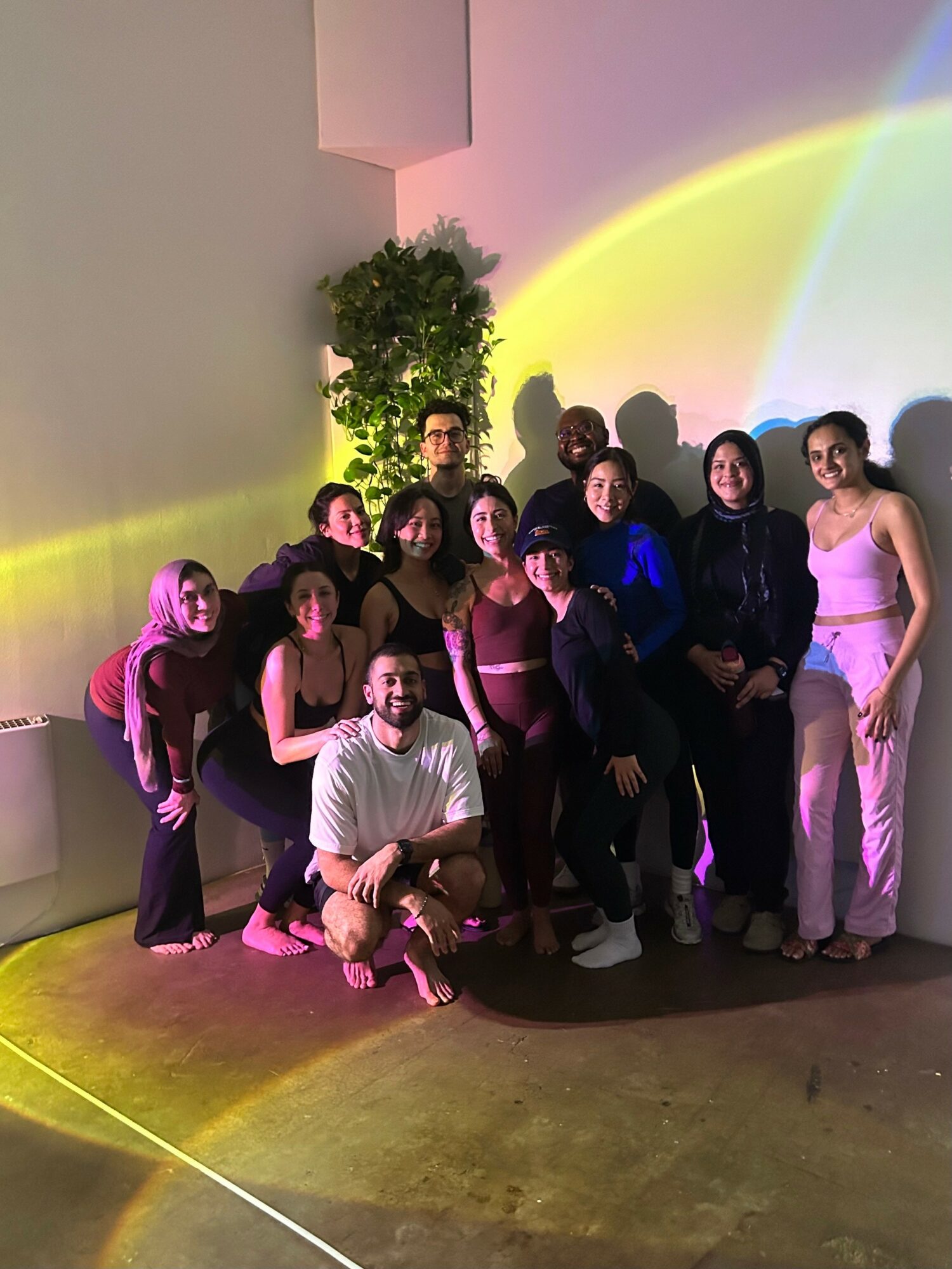 Group of ten people posing indoors with colorful lighting and a potted plant, some standing and some kneeling or sitting.