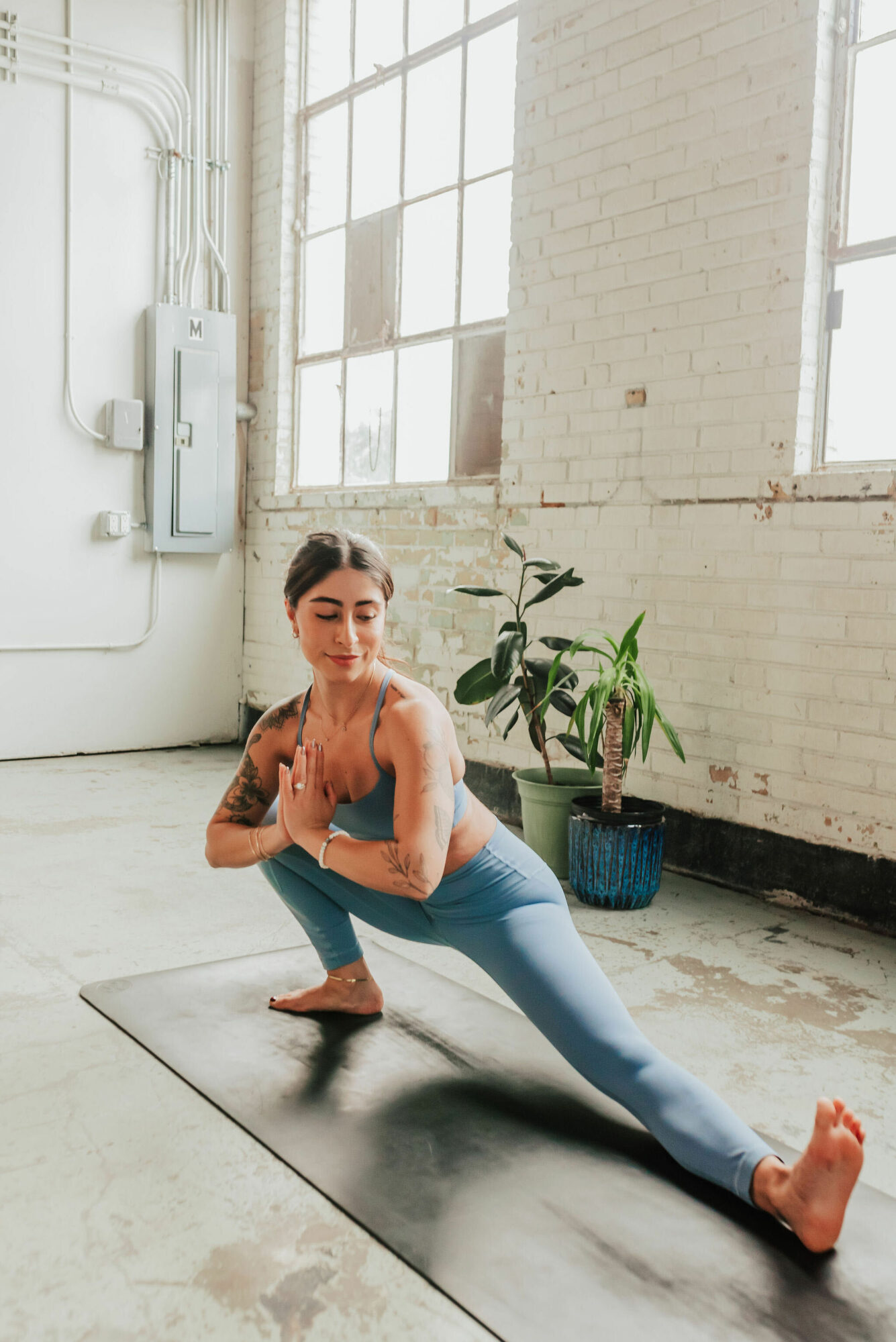Woman practicing yoga in a bright industrial-style room with large windows and a potted plant, in a wide lunge pose.