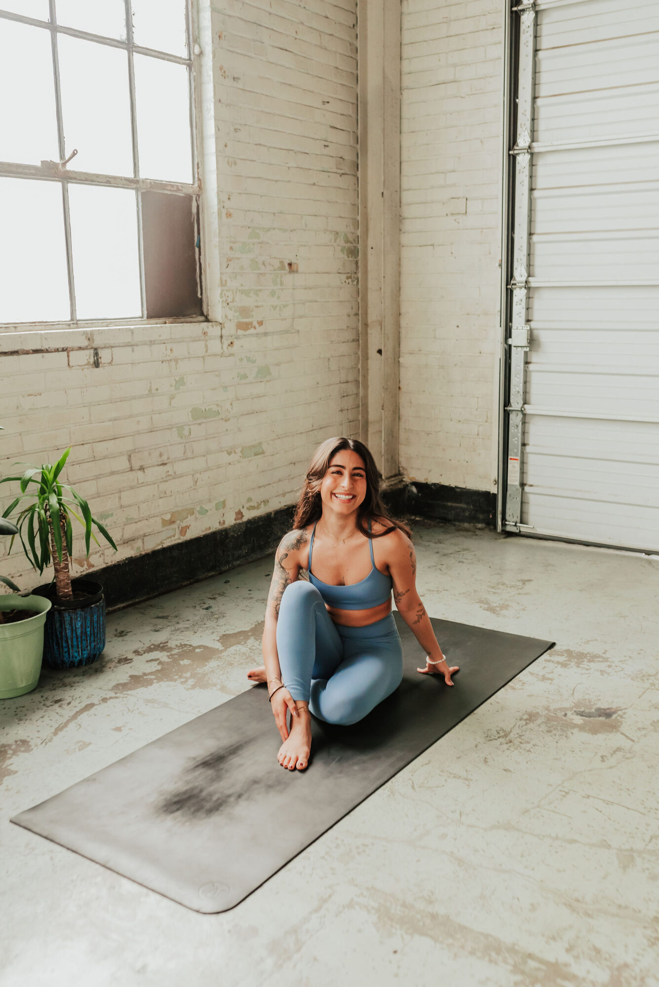 Woman practicing yoga on a mat in a bright industrial space with large window and potted plant.