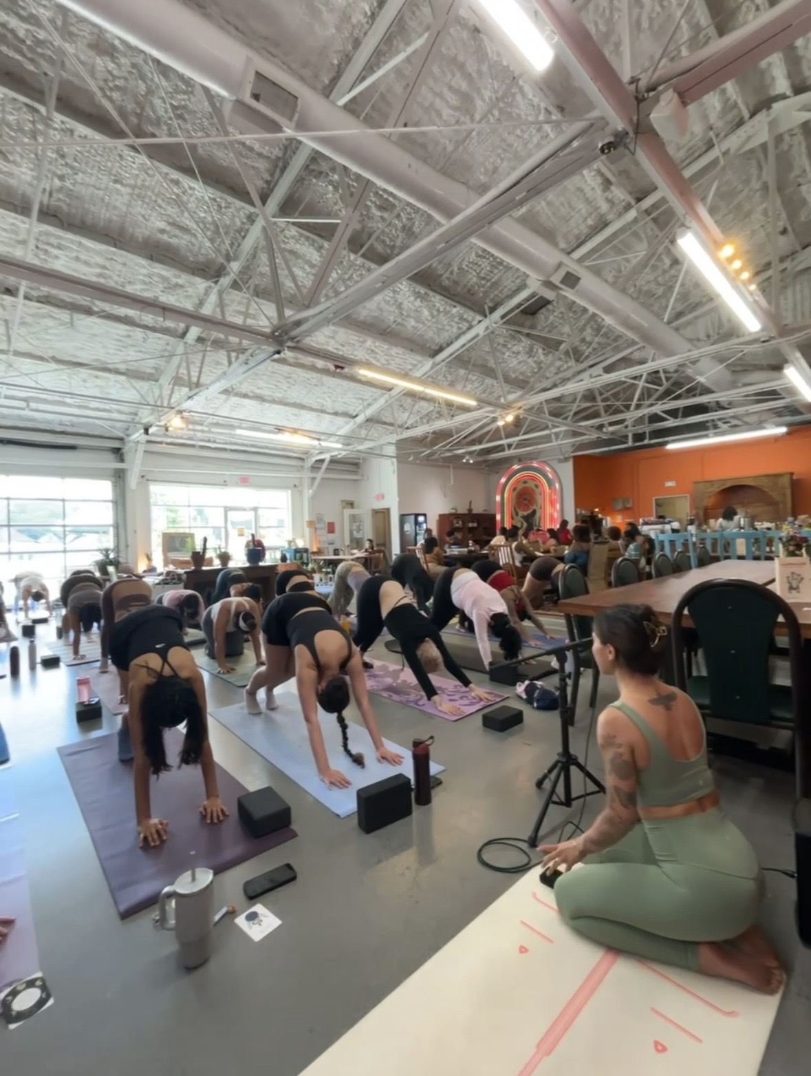 People practicing yoga in a spacious indoor studio with high ceilings and natural light, some in downward dog pose, instructor seated nearby.