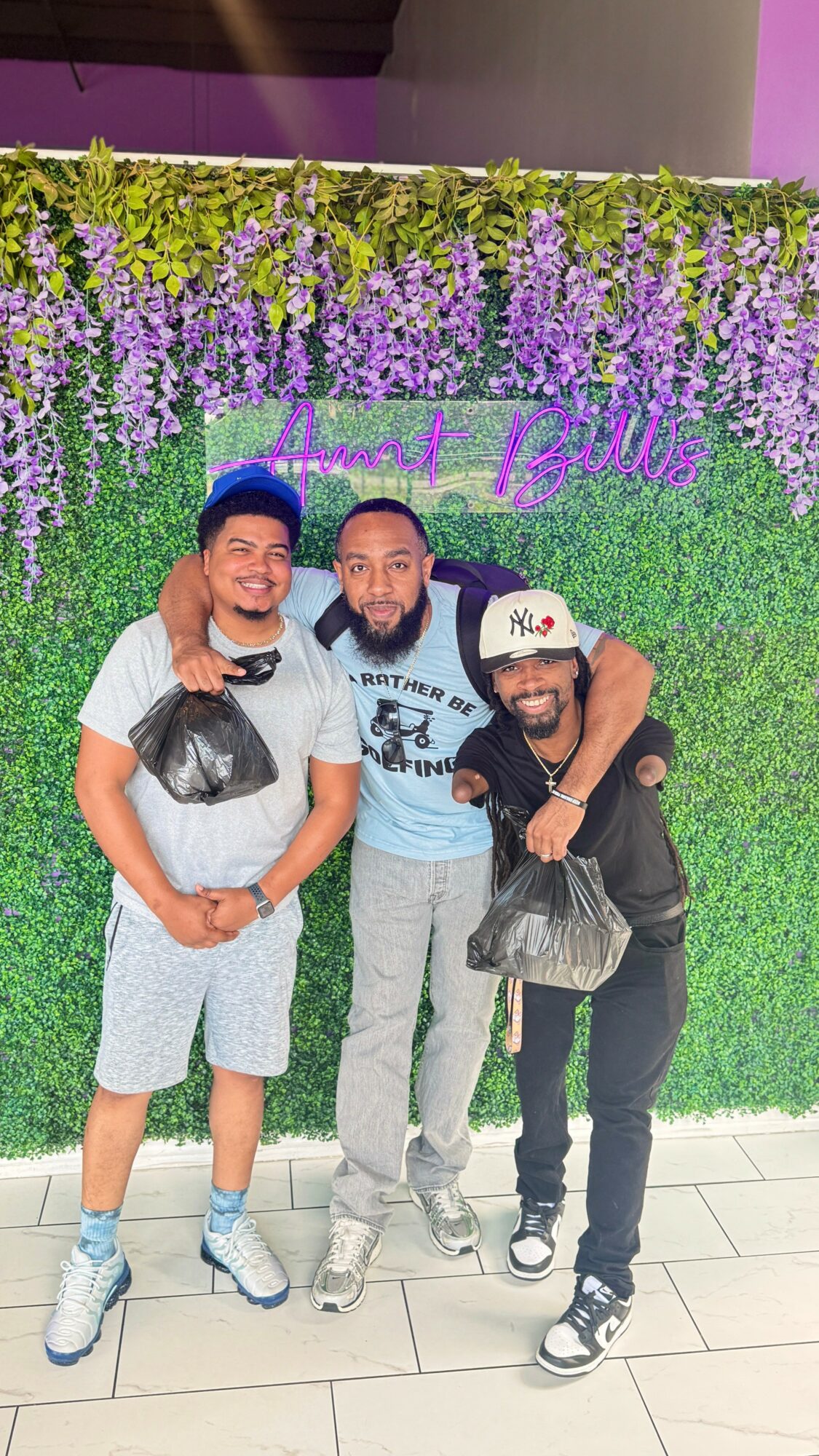 Three people standing in front of a green and purple floral backdrop, smiling and hugging each other.