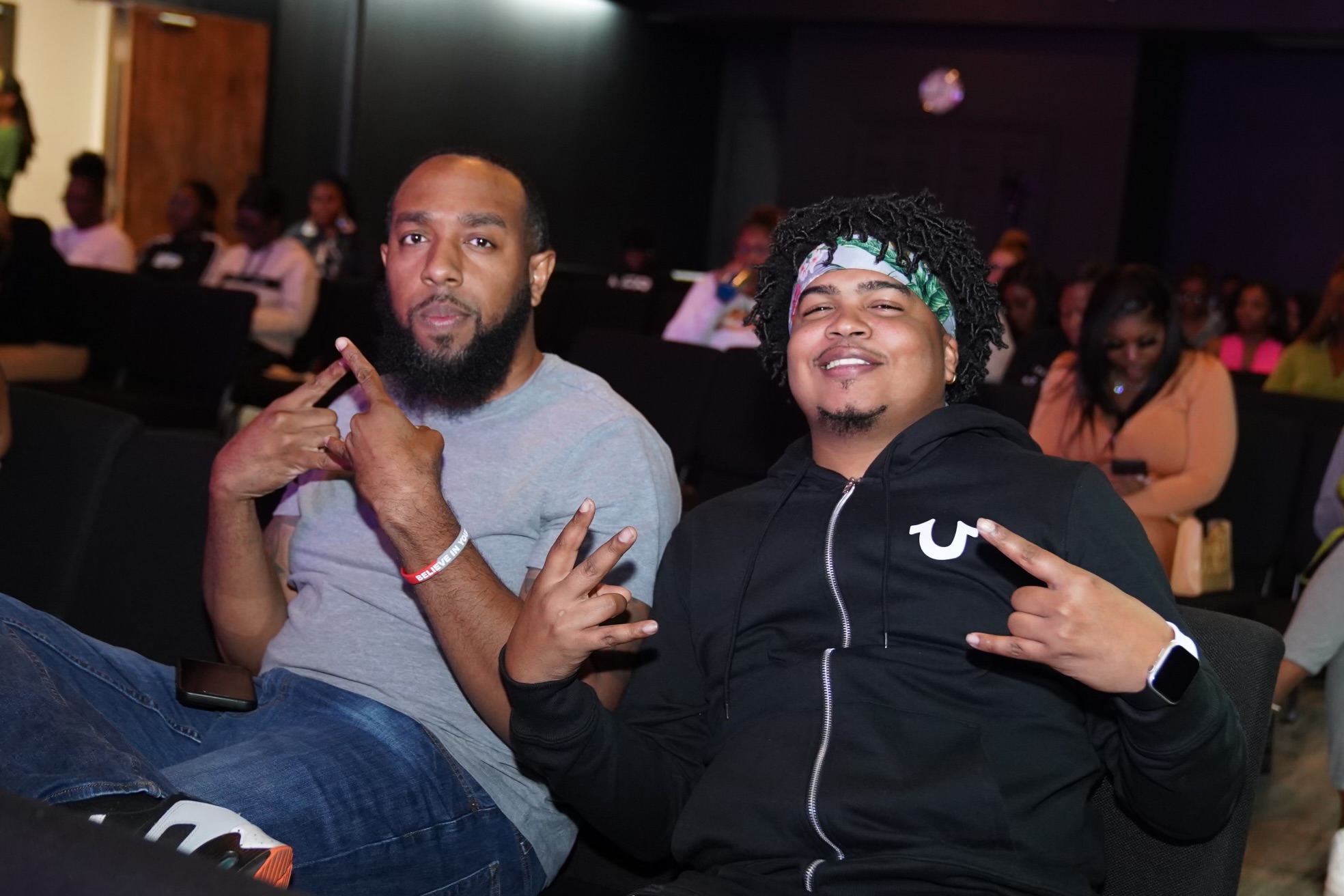 Two men sitting in a crowded indoor space, smiling and making hand gestures, with other people in the background.