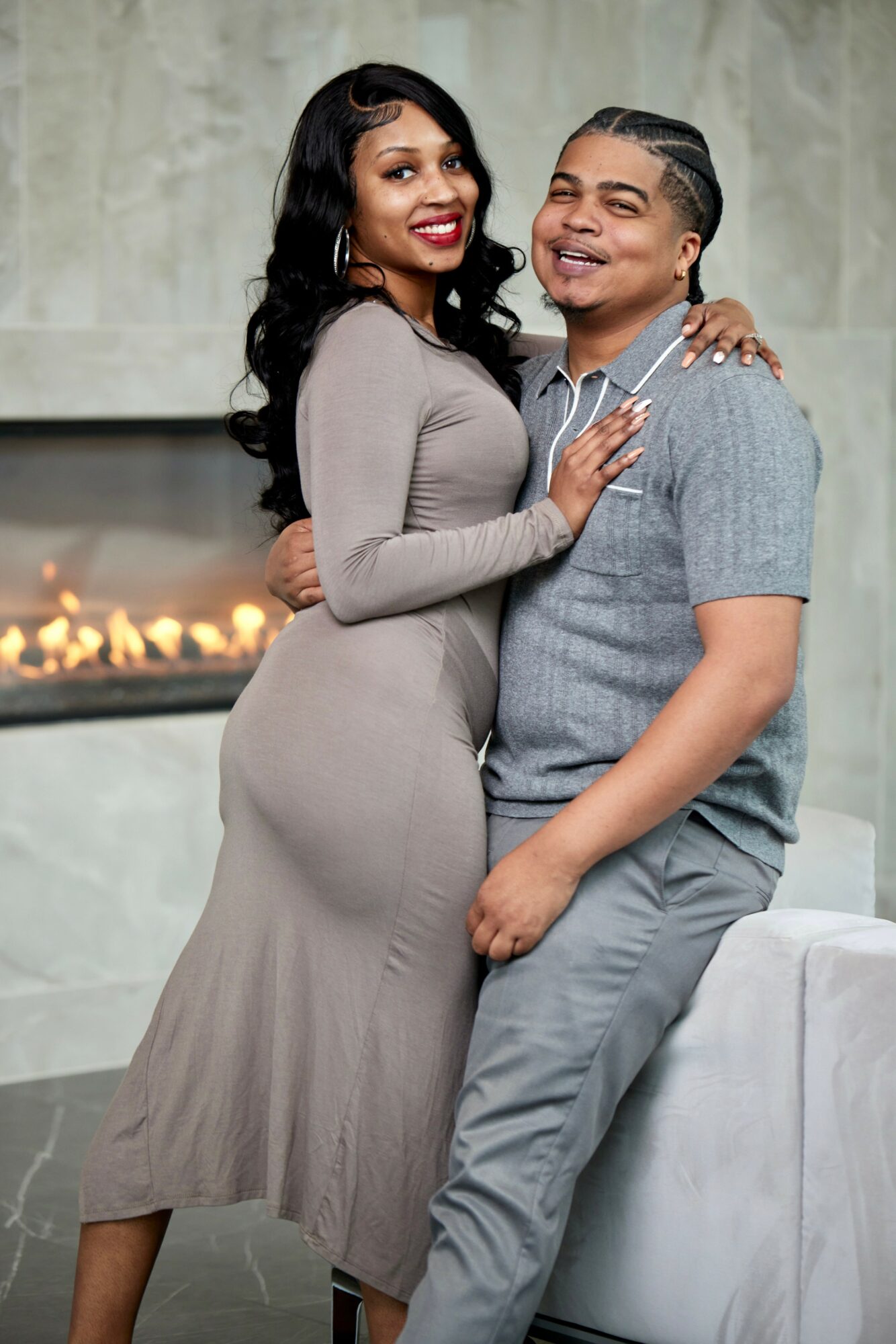 Two people smiling and hugging indoors, one wearing a long gray dress and the other in a gray shirt and pants.