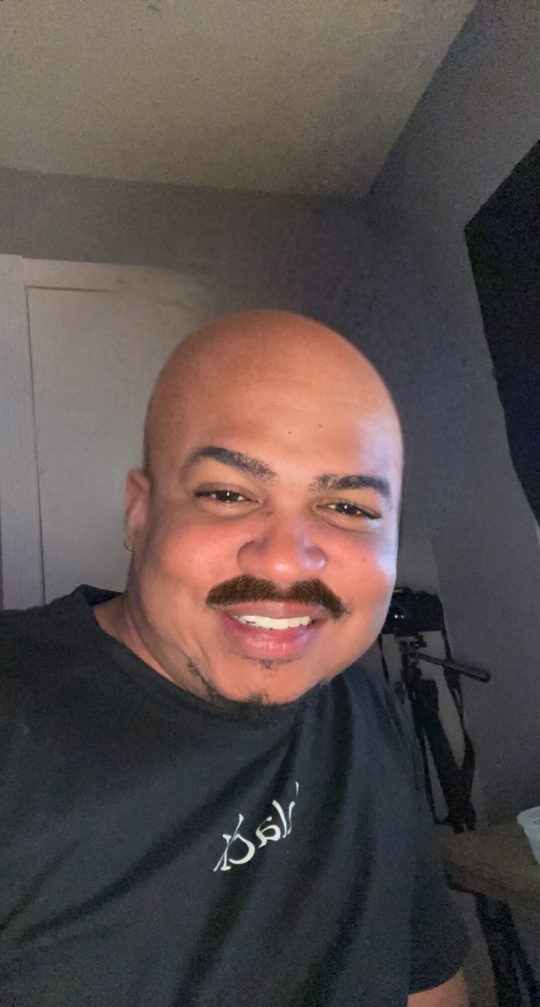 Smiling person with a bald head and mustache, wearing a black shirt, in a room with gray walls.