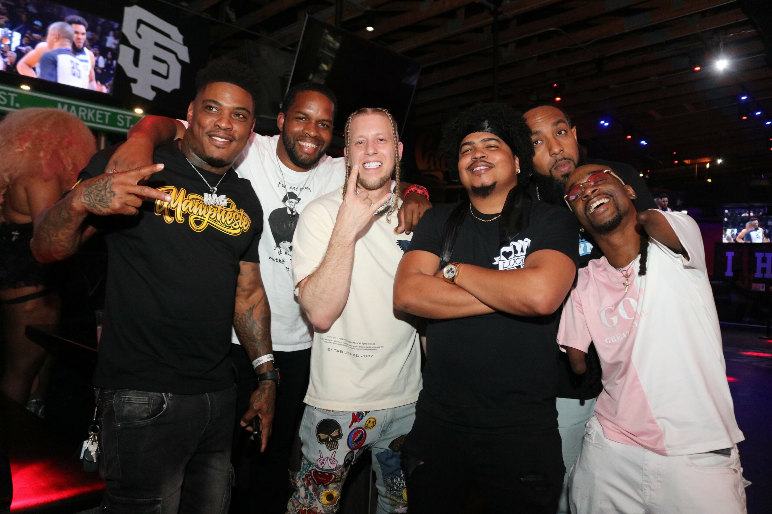Six men smiling and posing together in a dimly lit indoor setting, some making gestures, with a large screen in the background.