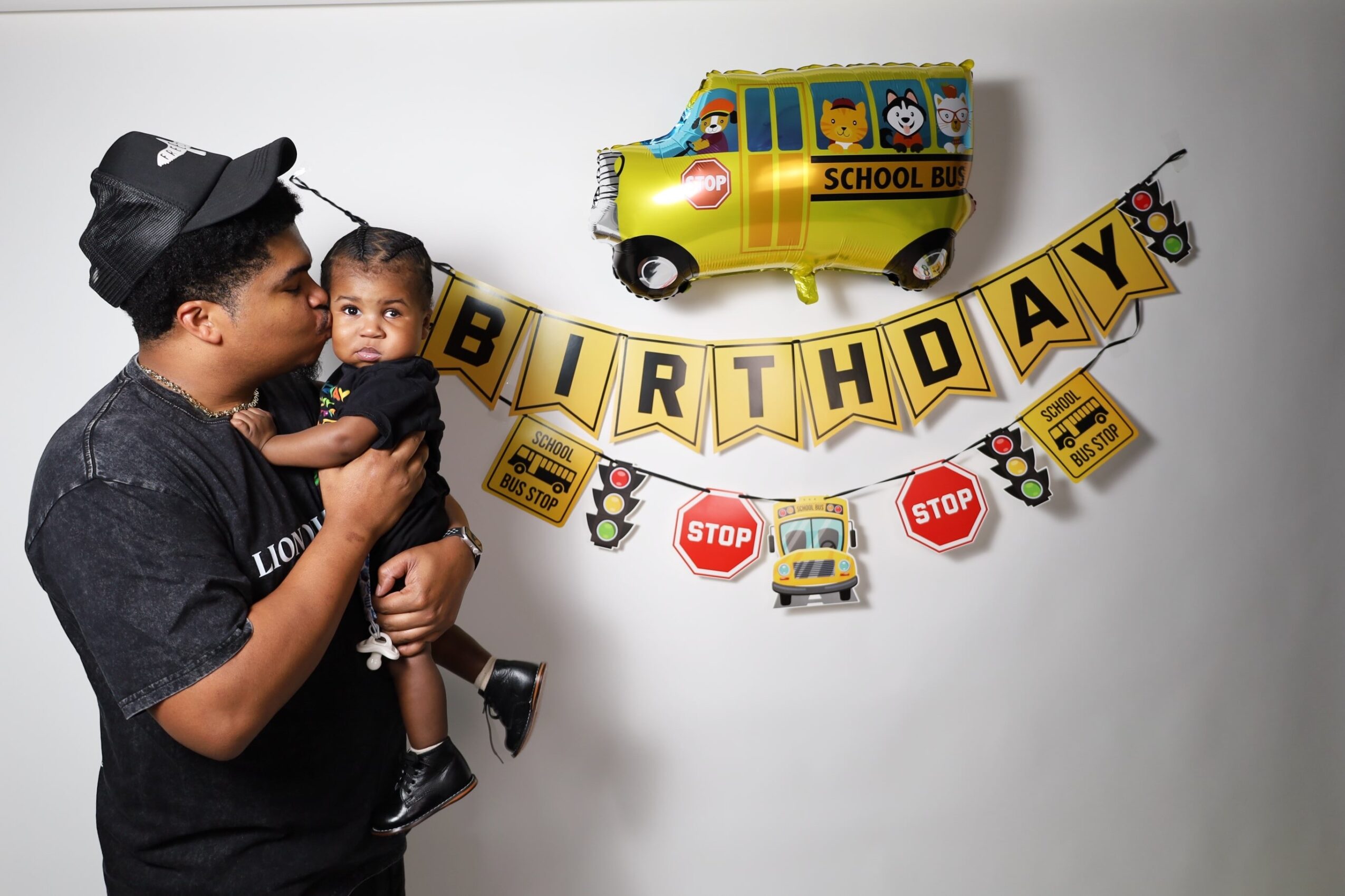 Adult holding a child in front of birthday decorations with a yellow school bus balloon and a banner with stop signs and a bus image.