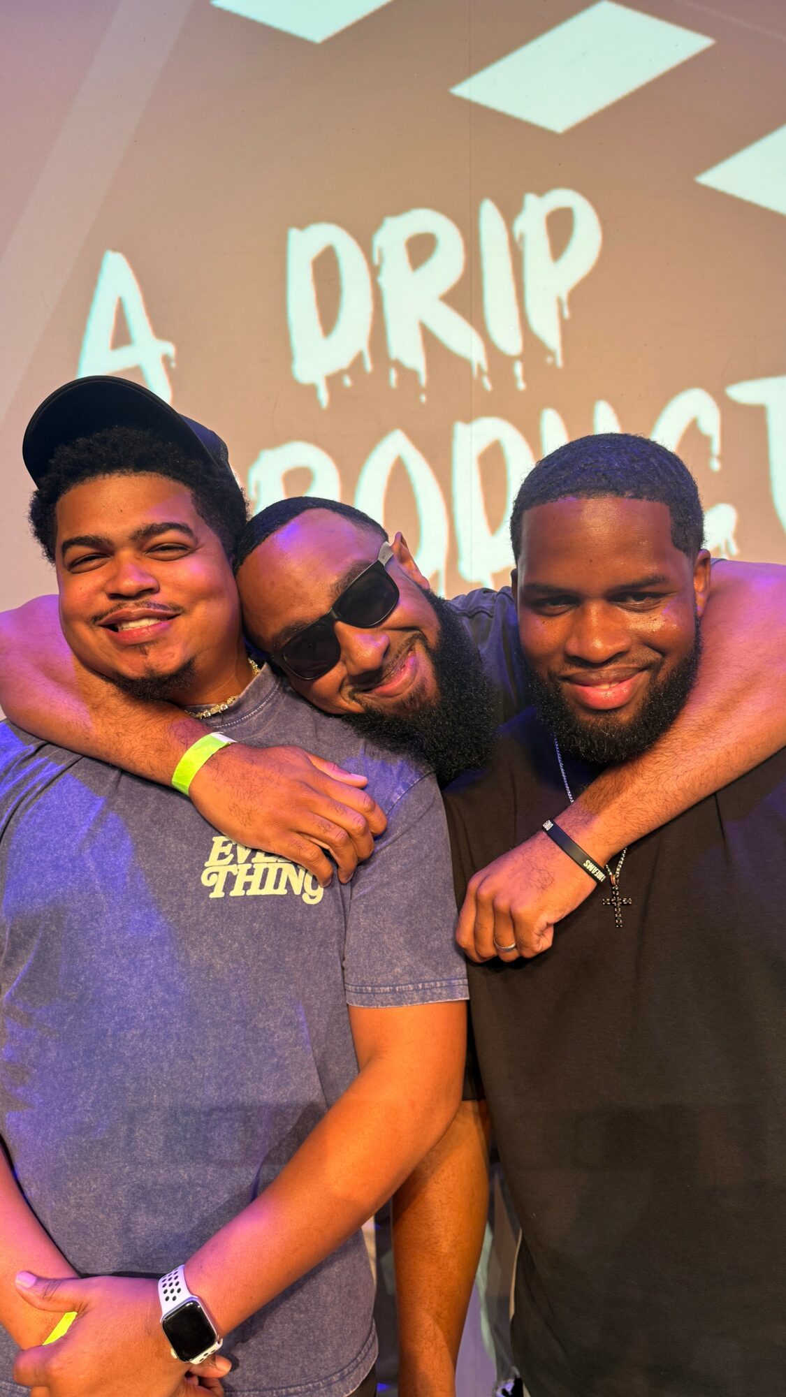 Three men smiling and hugging in front of a screen with text, one wearing sunglasses and a cap.