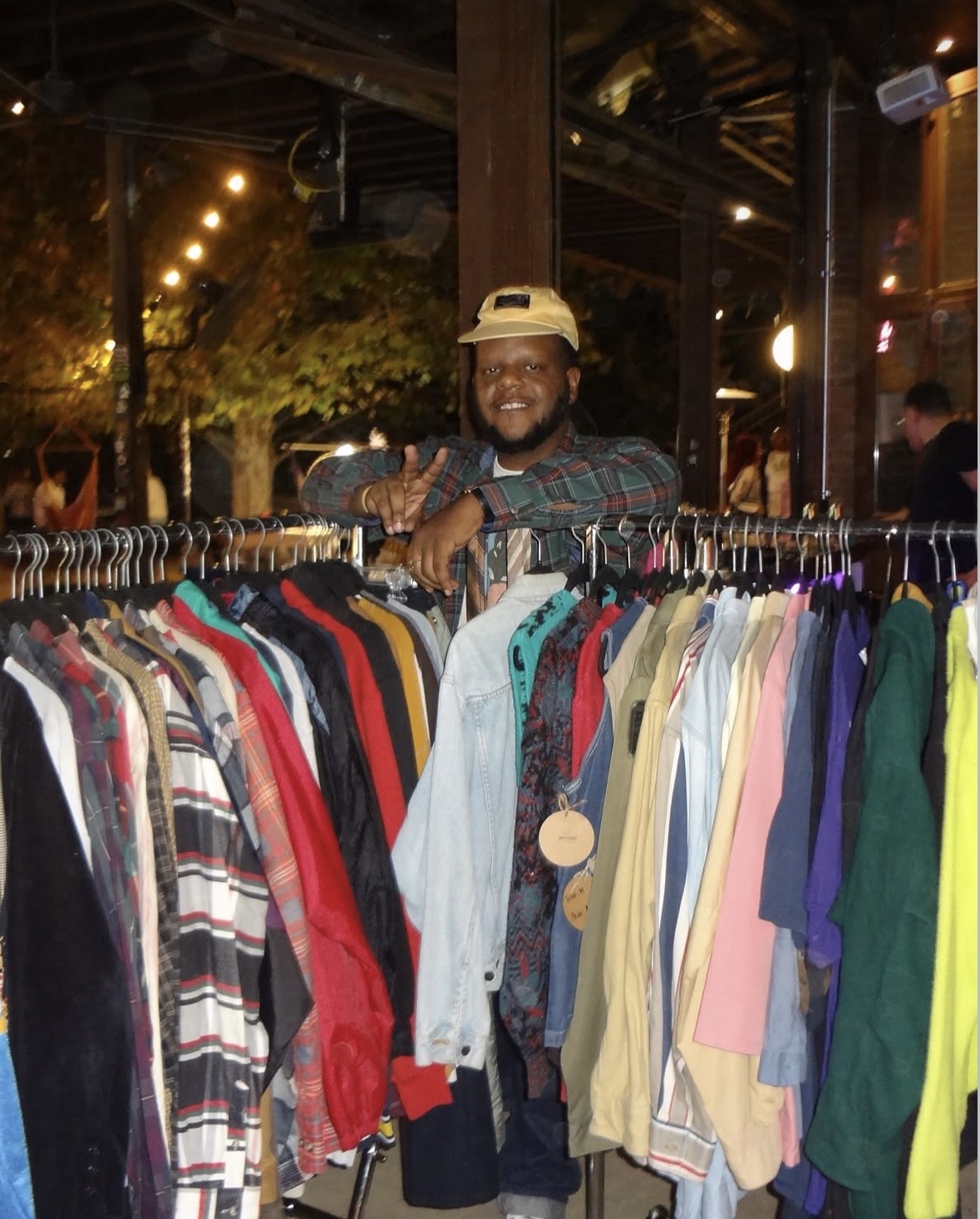 Man smiling behind a clothing rack outdoors at night, with string lights and trees in the background.