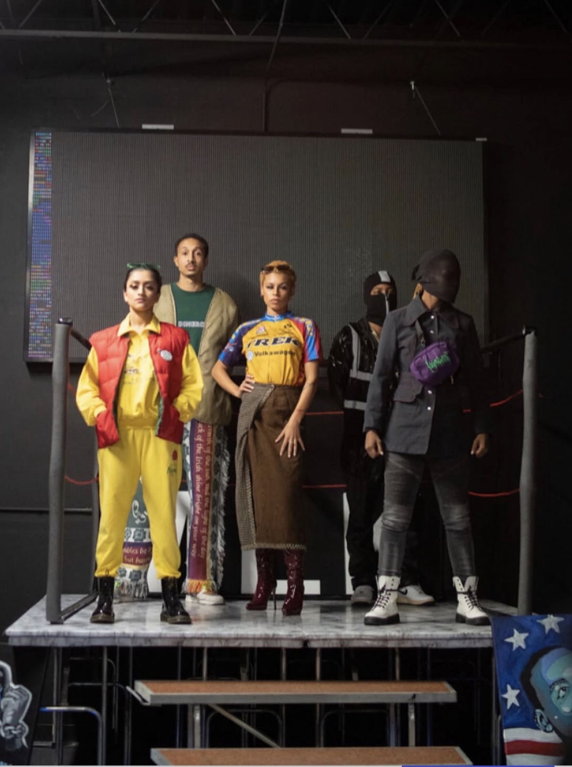 Five people stand on a stage with a large screen behind them, some facing forward and others turned away, in a dark setting.
