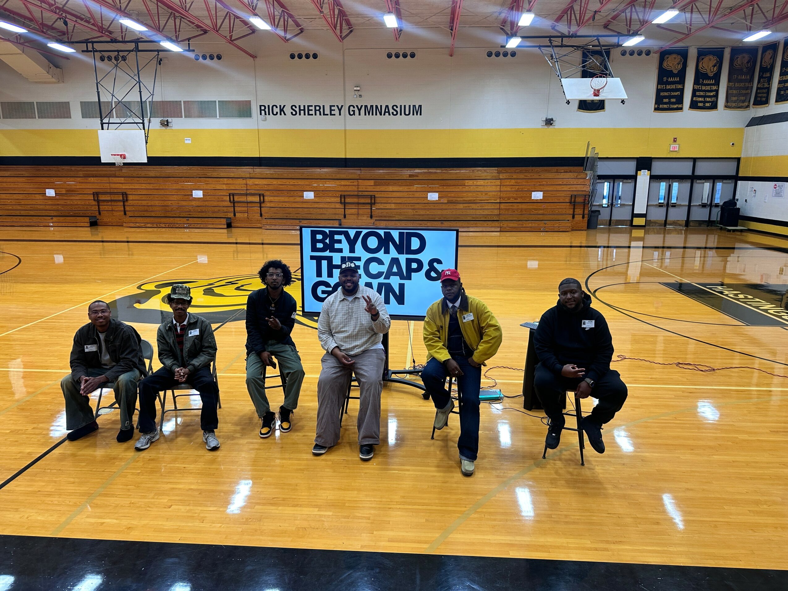 Six people sitting and standing in a gymnasium with a large screen behind them displaying 'BEYOND THE CAPS GYM'.