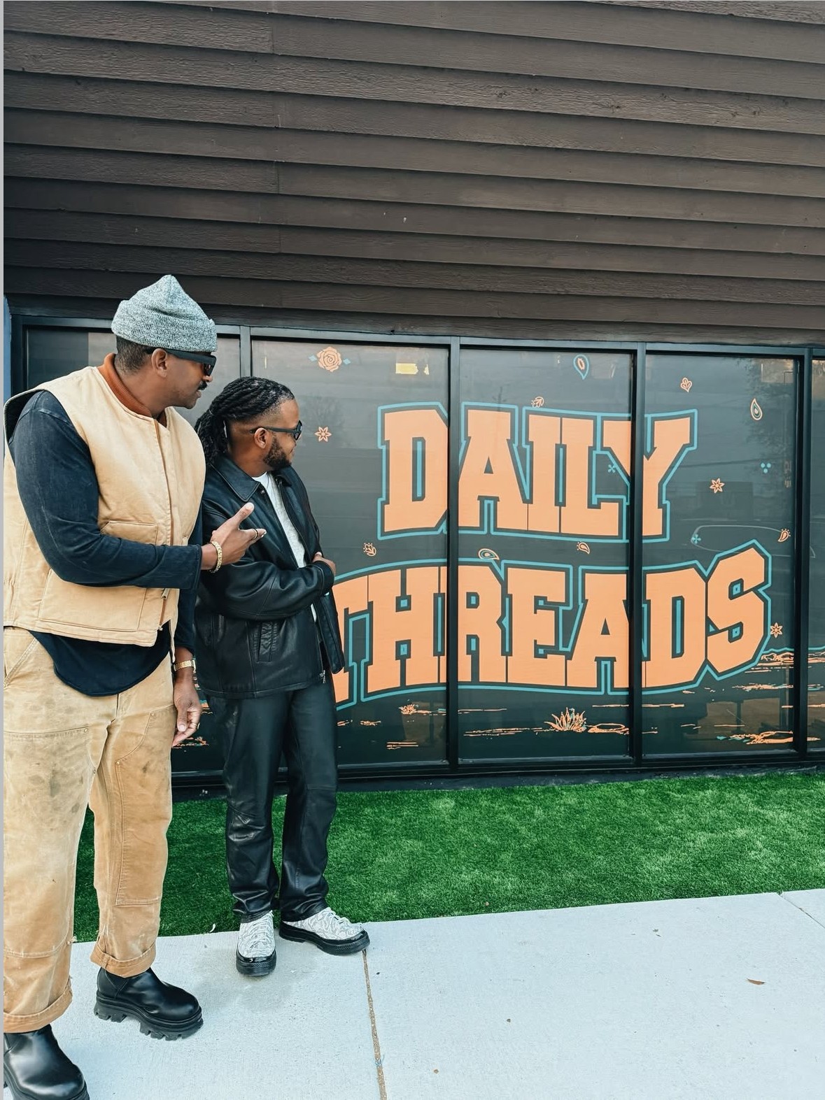 Two people standing outside near a sign that reads 'Daily Threads,' with a dark wooden wall behind them.