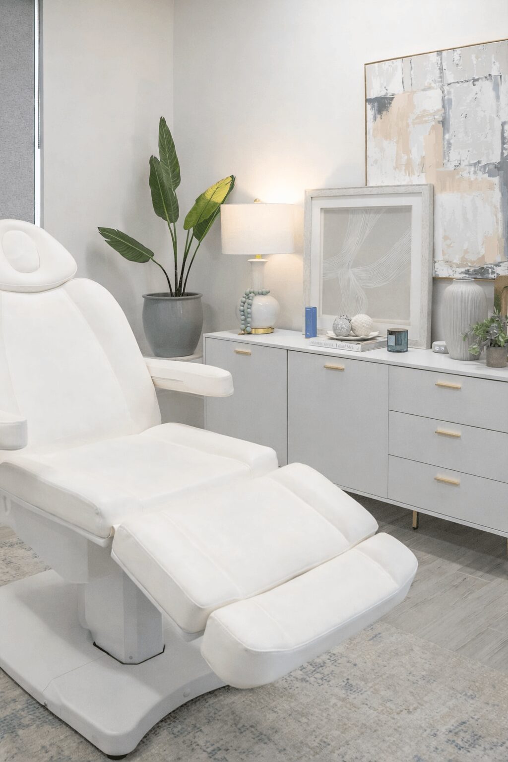 Medical examination chair in a room with a plant, lamp, and cabinet, all in neutral tones.