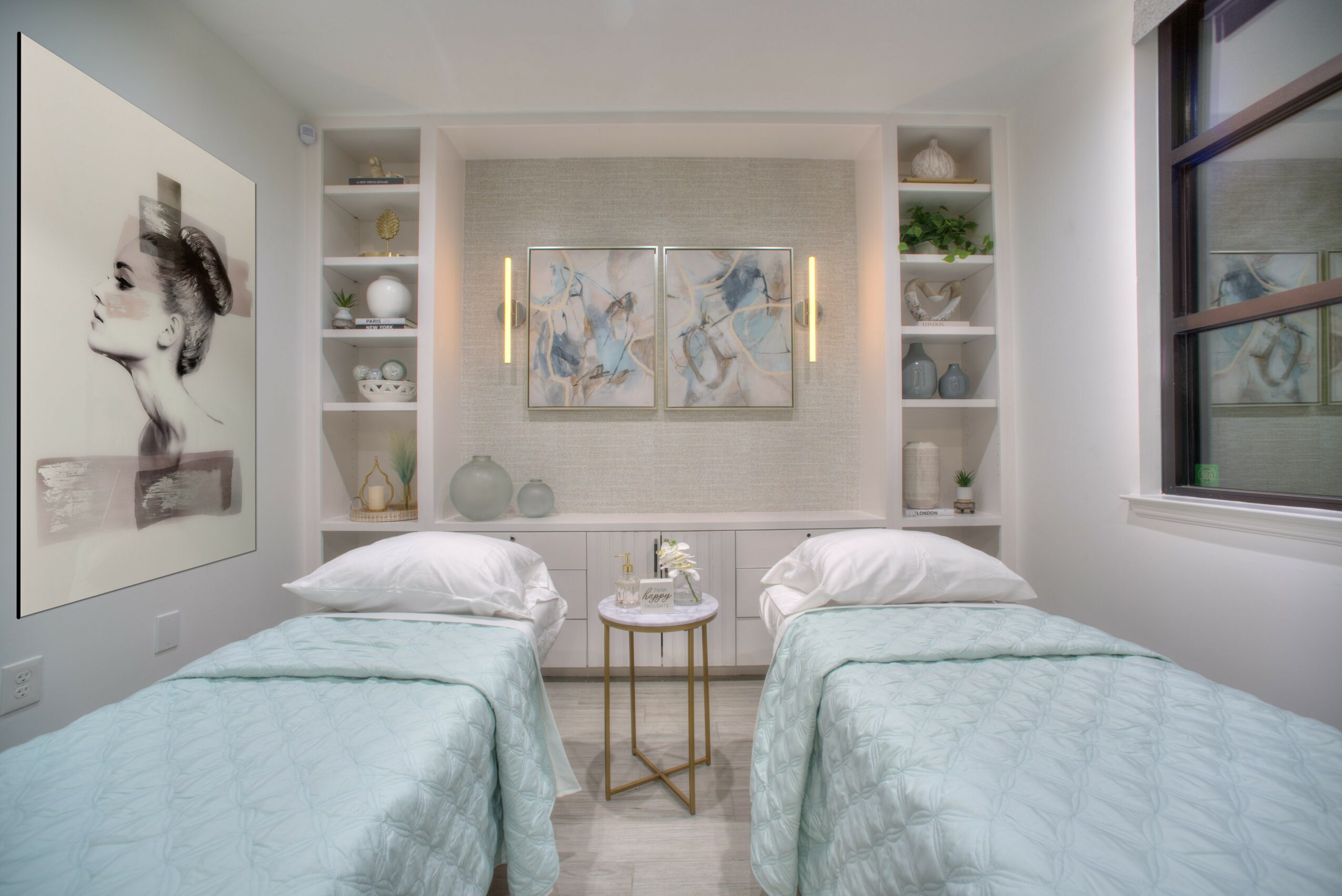 Two beds with light blue bedding and pillows, a small round table between them, wall art, and built-in shelves with decor, window on right.