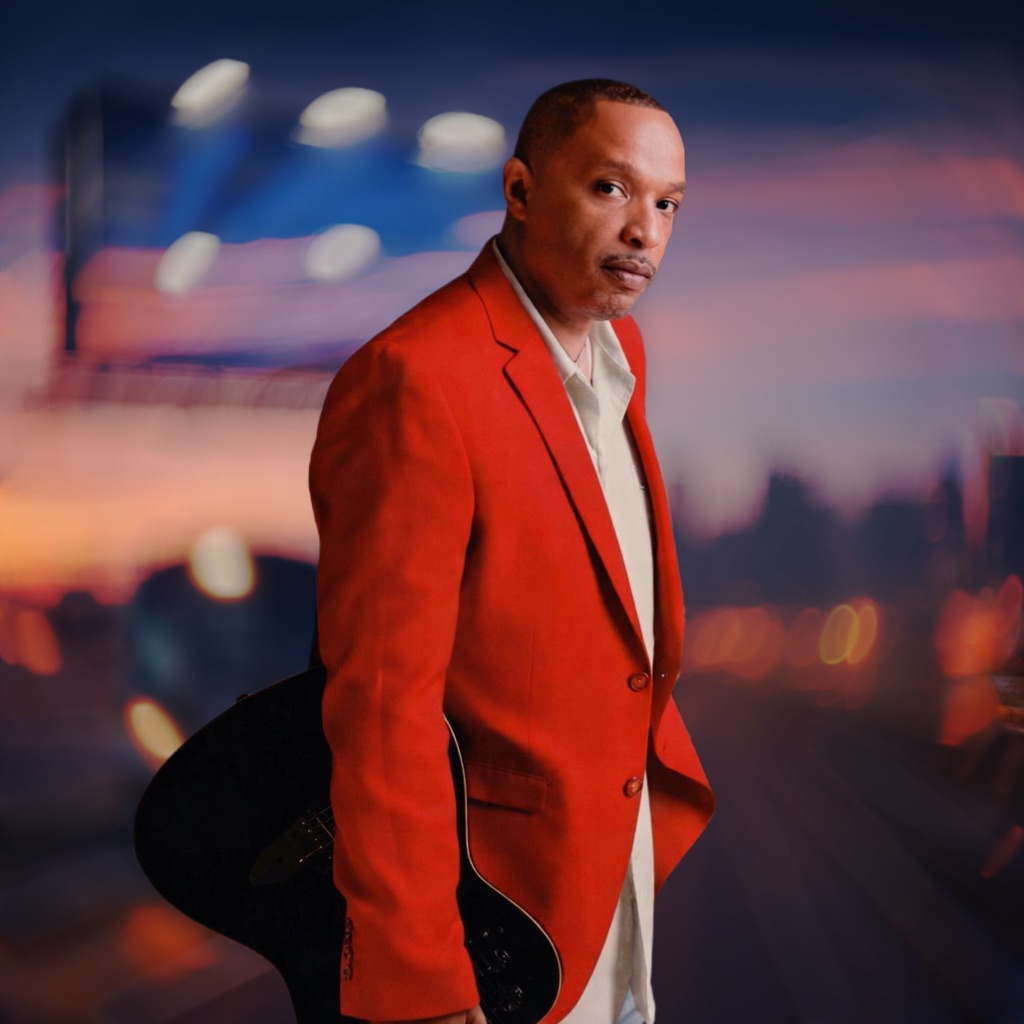 Man in red blazer standing outdoors with cityscape and sunset in background.