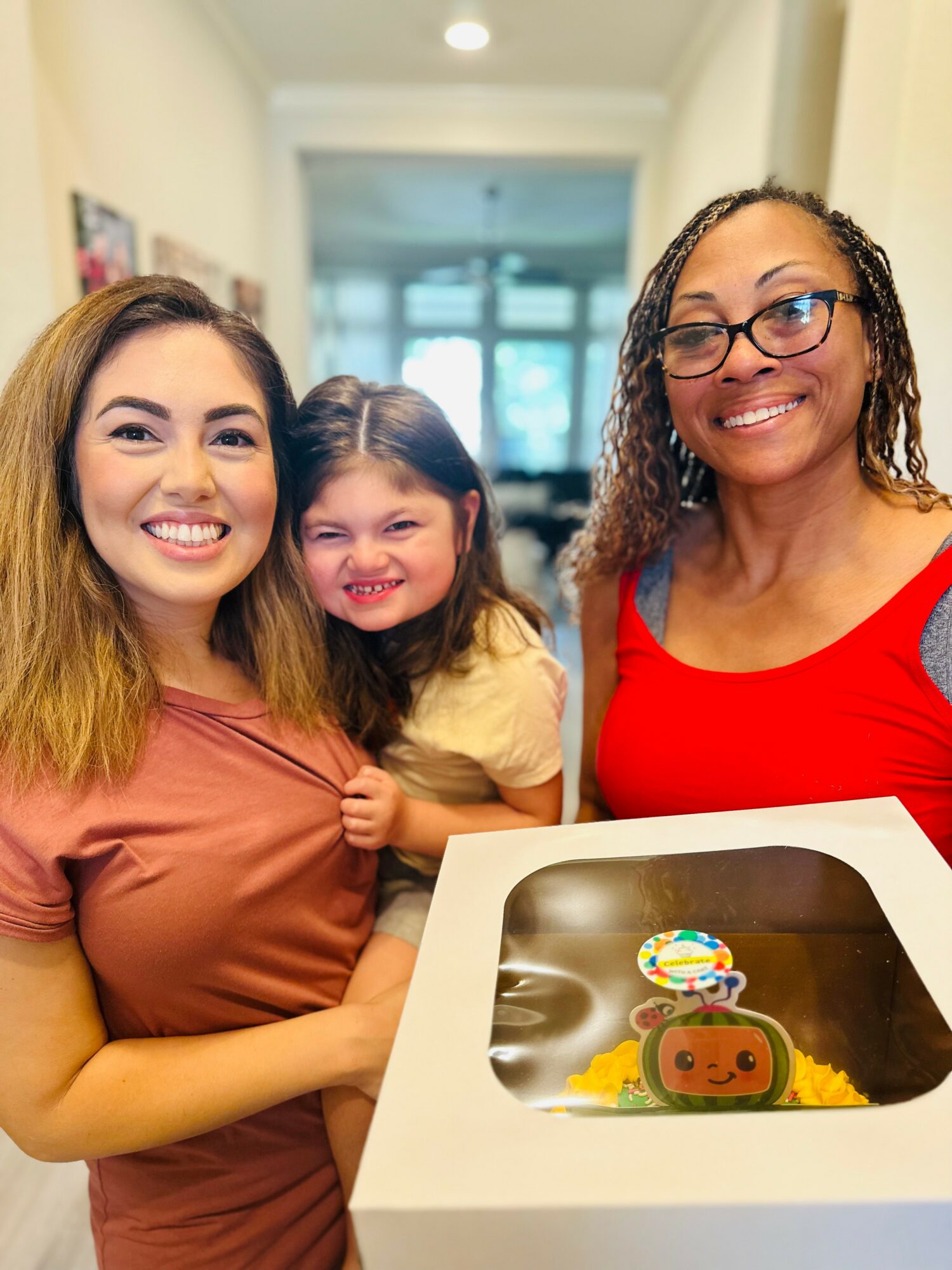 Three smiling women and a girl pose indoors, with a cake in a box in front of them.