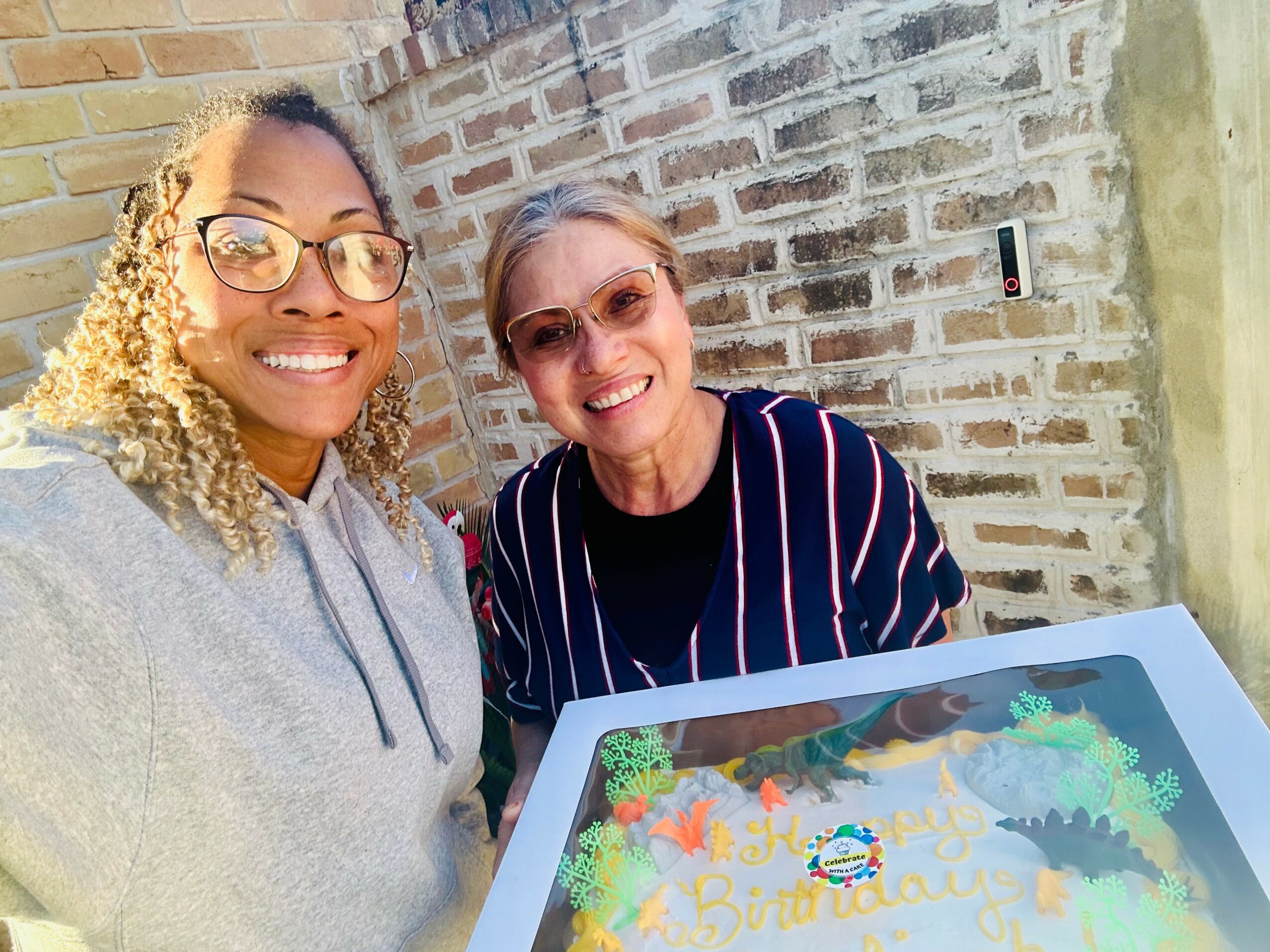 Two women smiling outdoors near a brick wall, taking a selfie with a birthday cake in front of them.