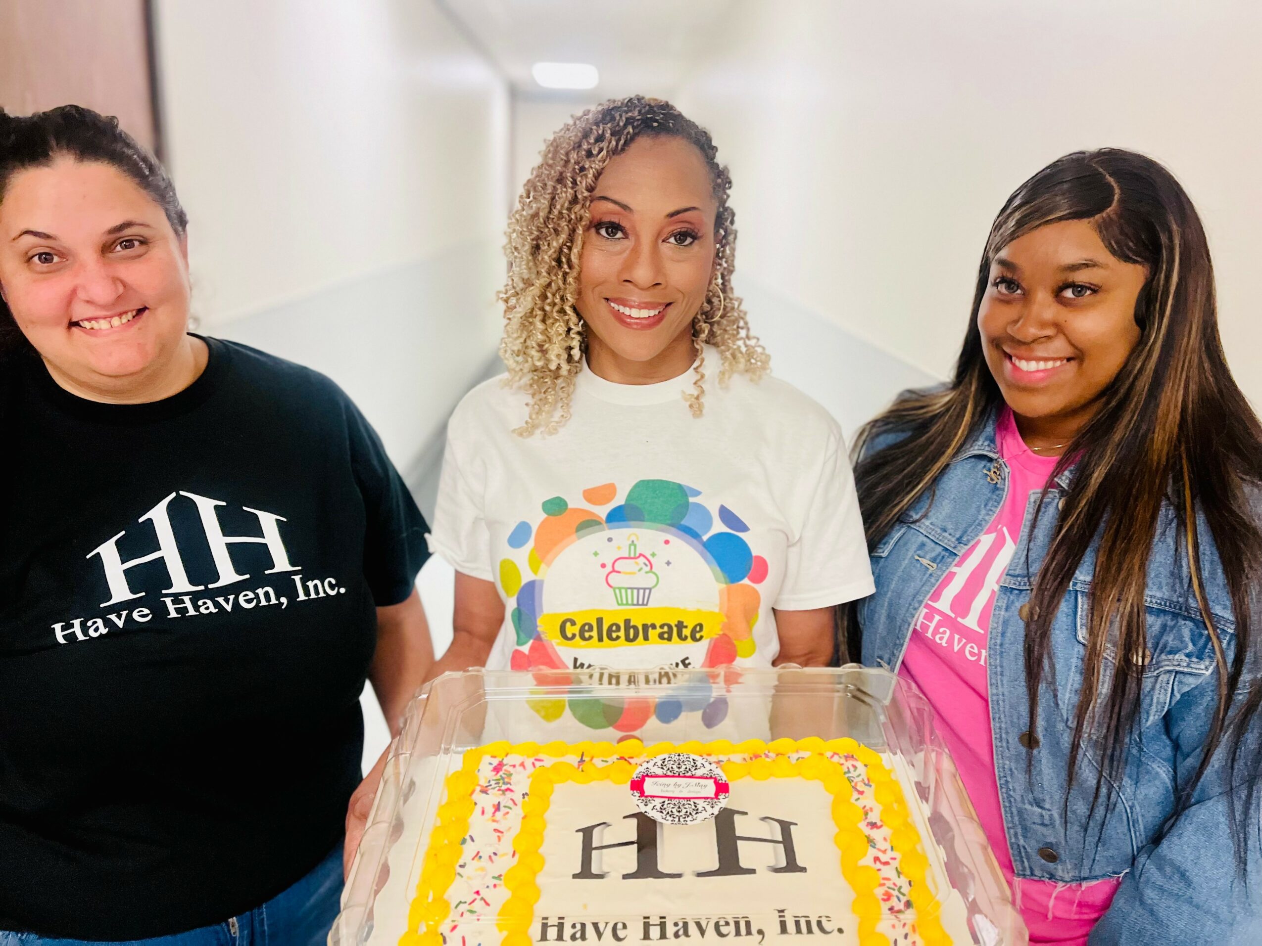 Three women standing behind a cake with a white background, smiling, celebrating, with a cake that has a yellow border and 'HH' written on it.