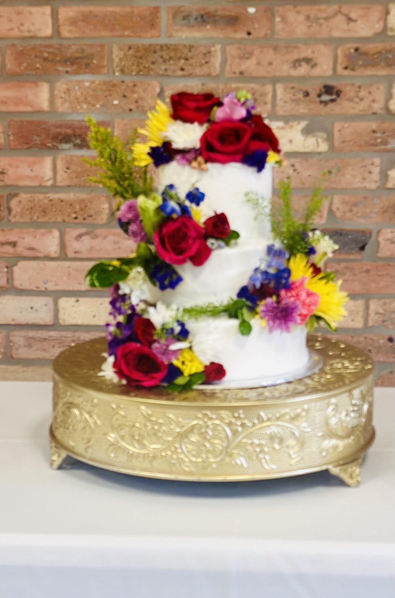 Two-tiered white cake decorated with colorful flowers, on a gold ornate stand, against a brick wall background.