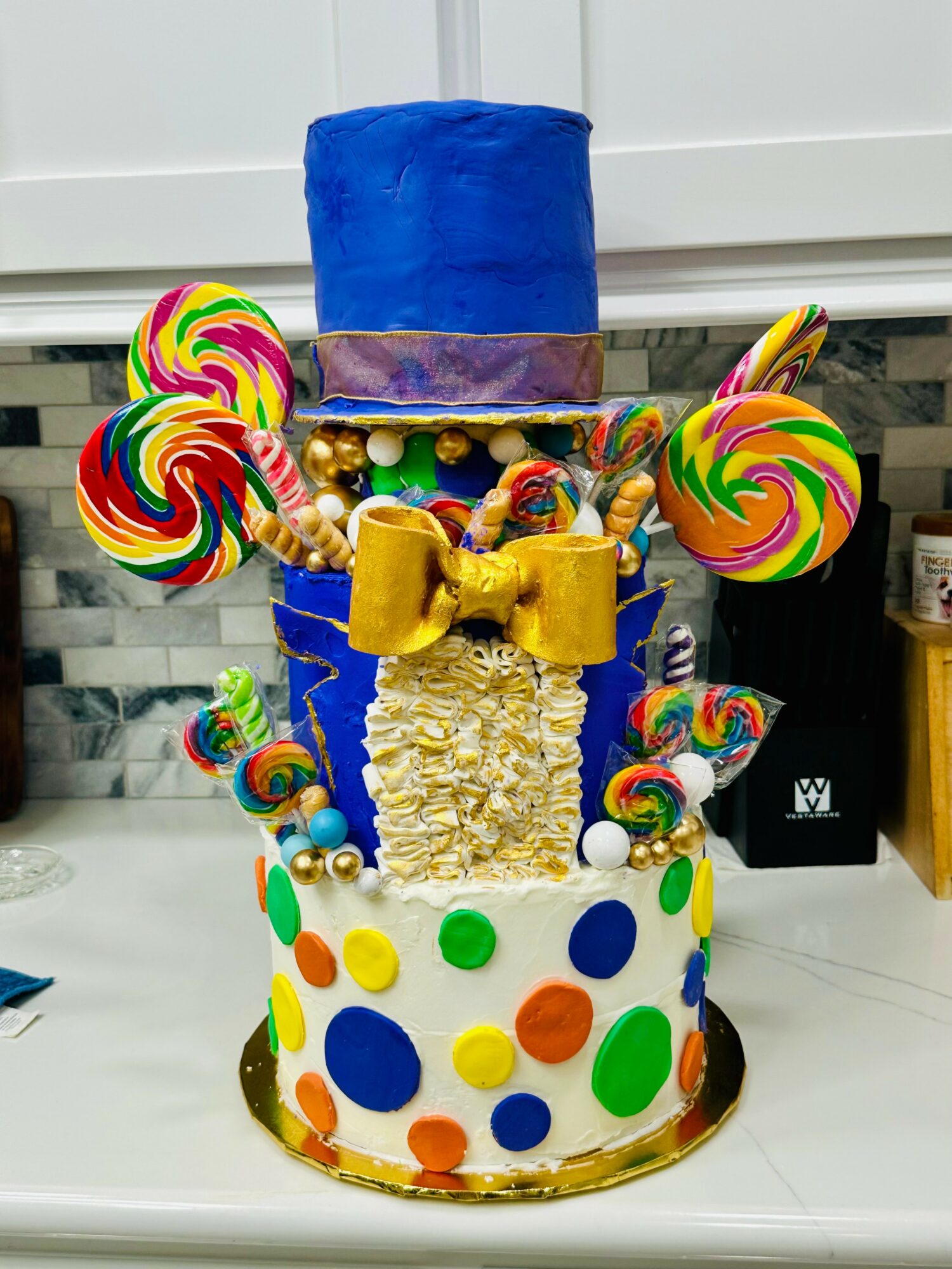 Colorful, multi-tiered cake with polka dots, lollipops, and a blue top hat decoration, set on a white surface.