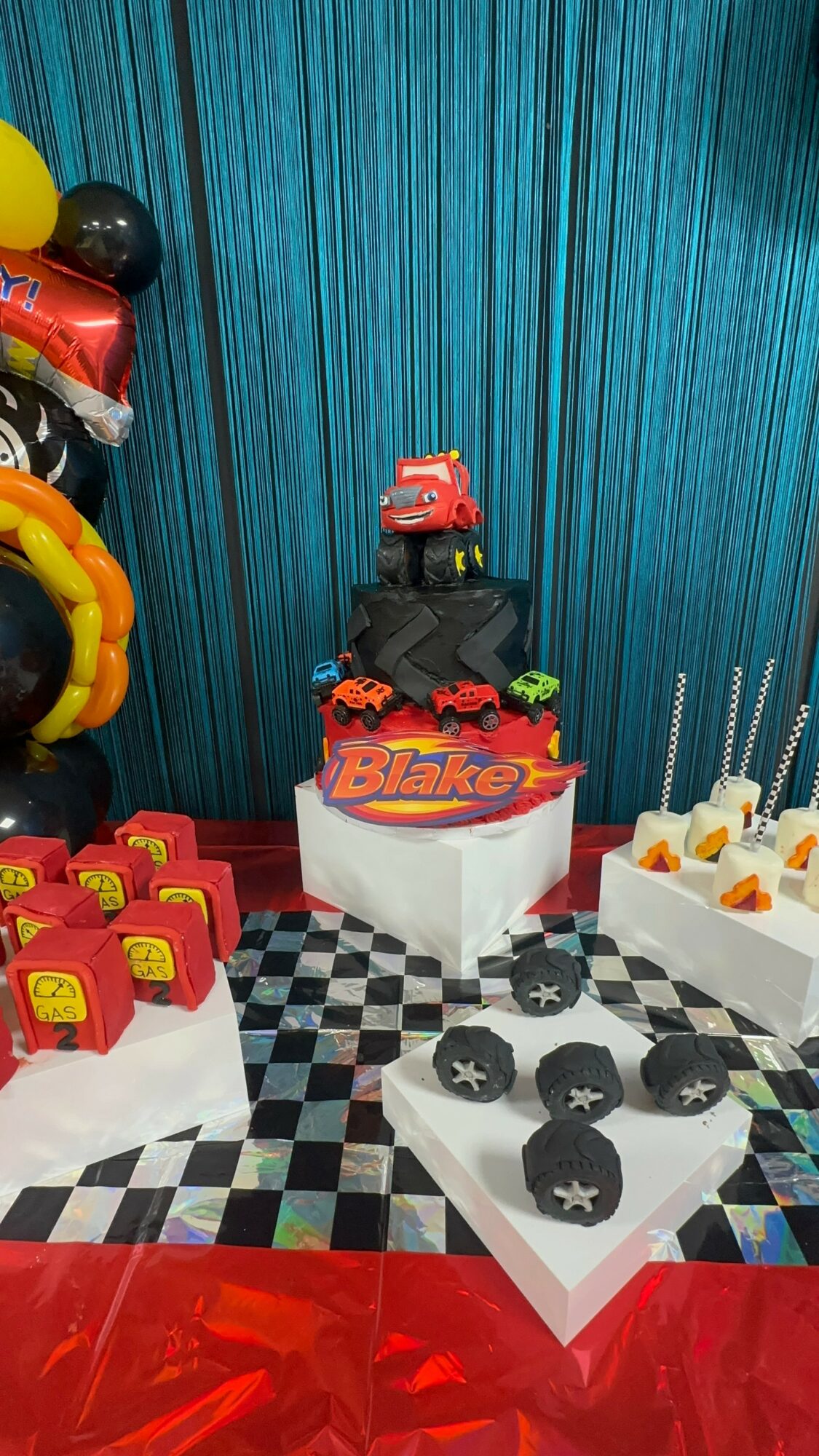 Party table with themed cake, cupcakes, and decorations featuring cars and racing elements, on a checkered tablecloth.
