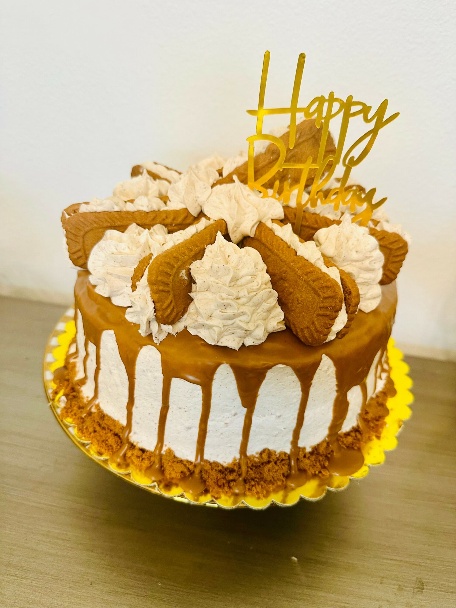 Round birthday cake with caramel drizzle, whipped cream, and waffle pieces, topped with a gold 'Happy Birthday' topper, on a gold base.