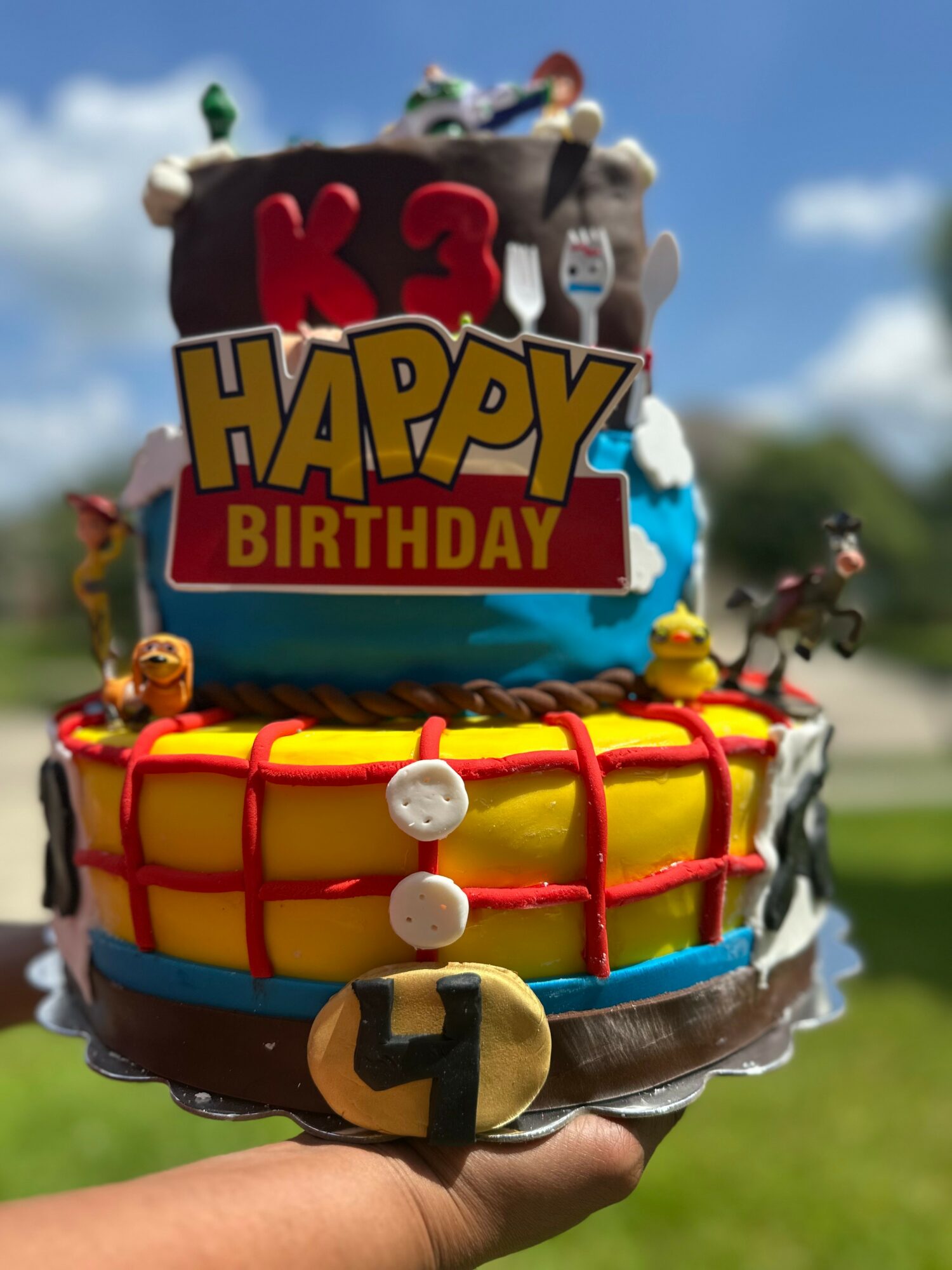 Colorful birthday cake with multiple layers, decorations, and a 'Happy Birthday' sign, held outdoors against a blue sky.