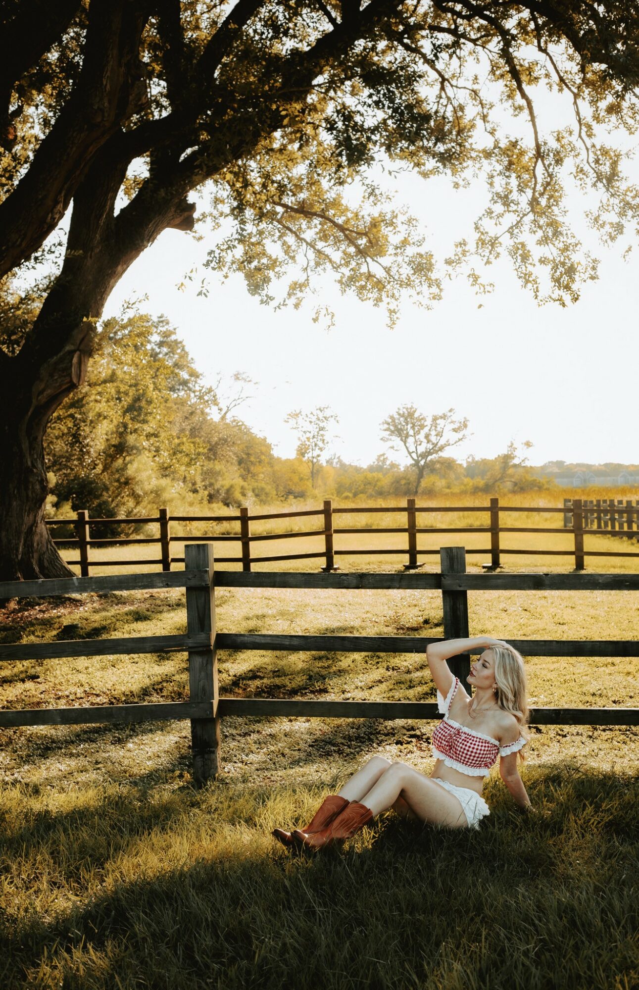Child sitting on grass near a wooden fence under a large tree in a sunny field.