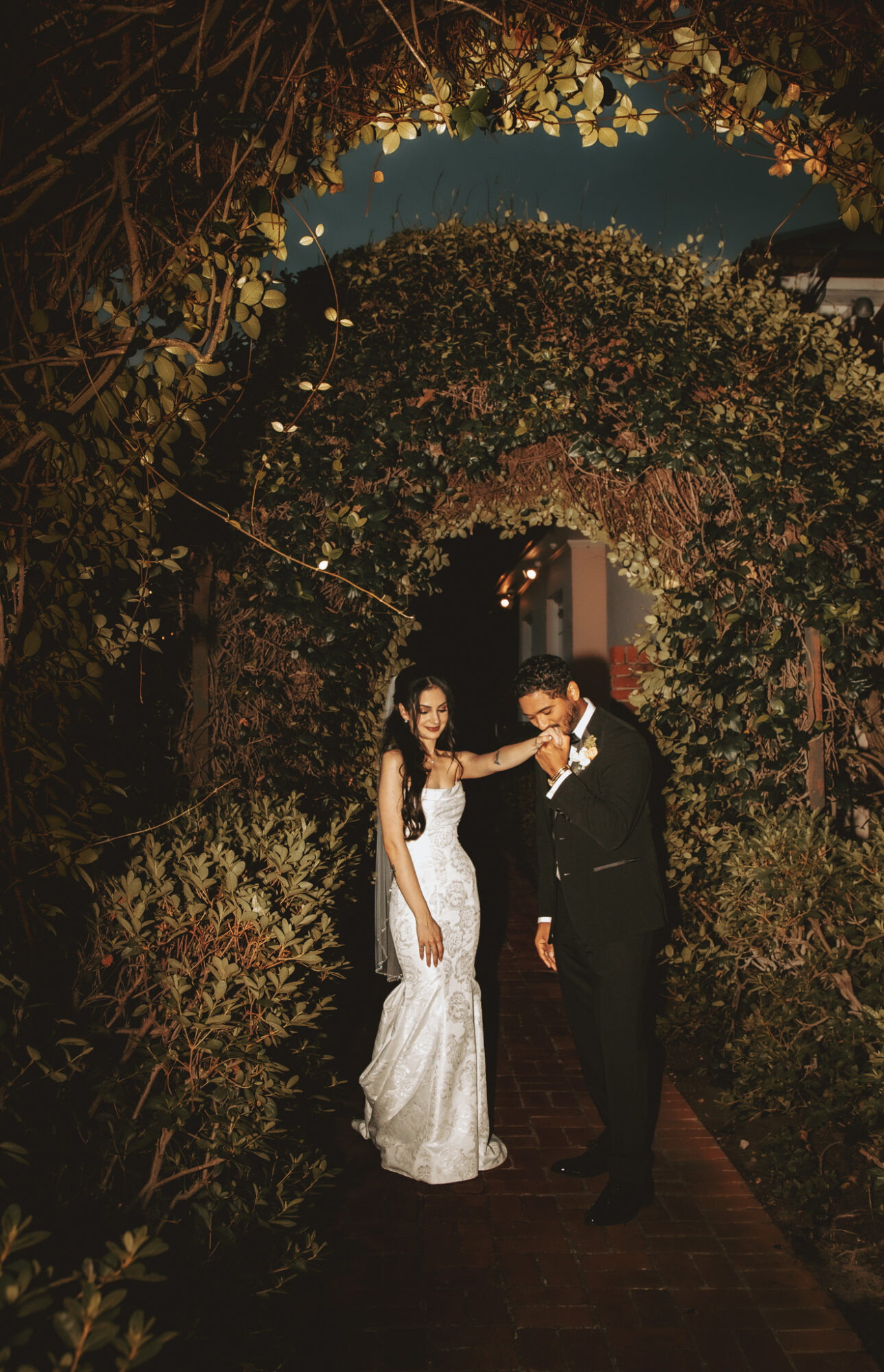 A bride and groom stand under an archway of flowers and greenery at night, holding hands and smiling.