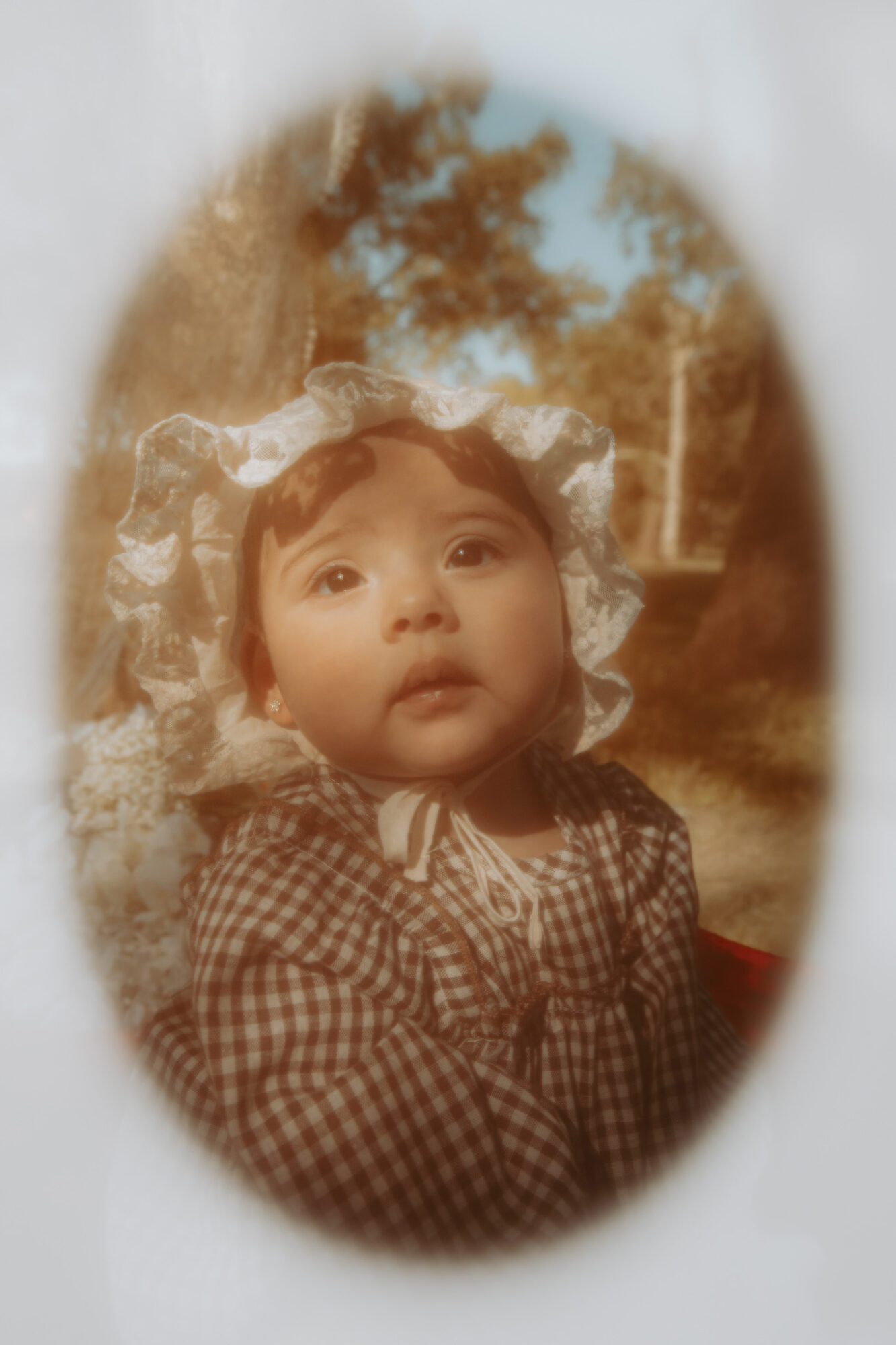 Child with a checkered shirt and bow headband outdoors, looking up, surrounded by trees and sky.