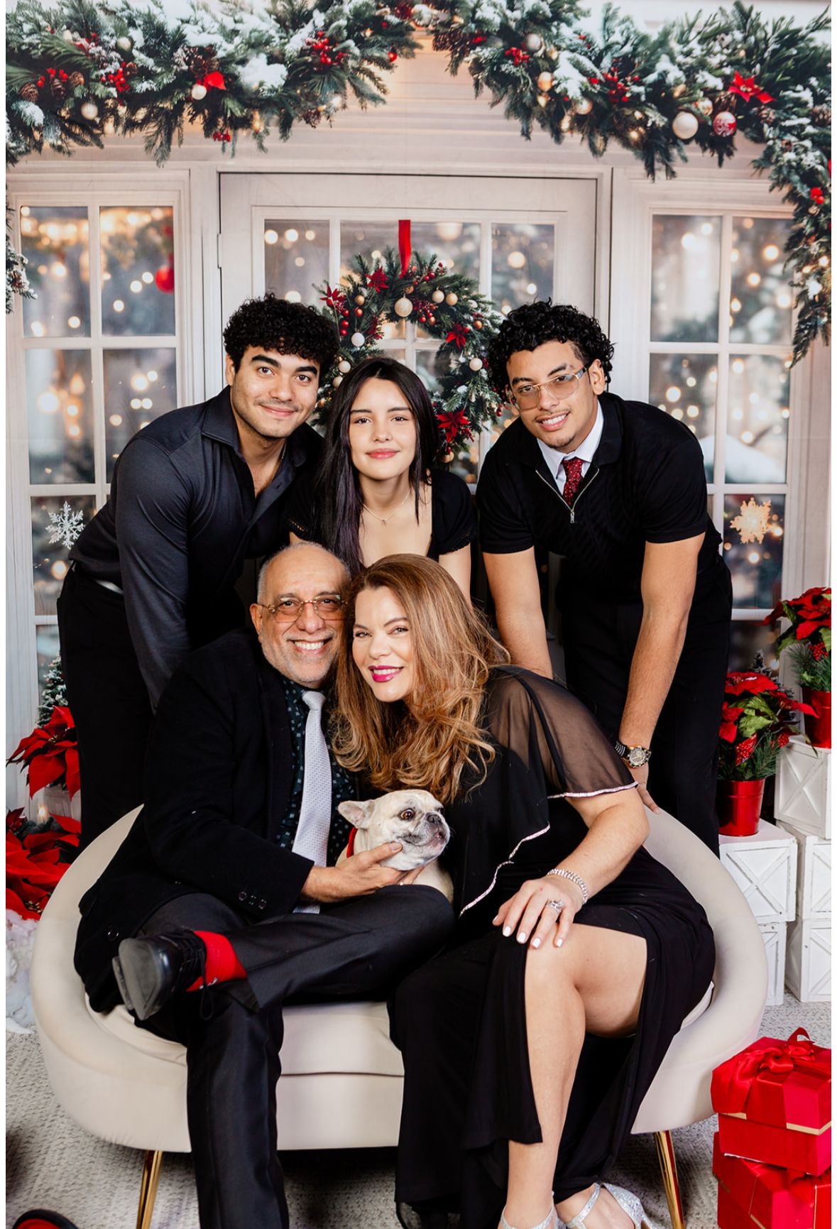 Group of five people sitting and standing in front of a decorated Christmas backdrop, smiling, with a red gift box nearby.
