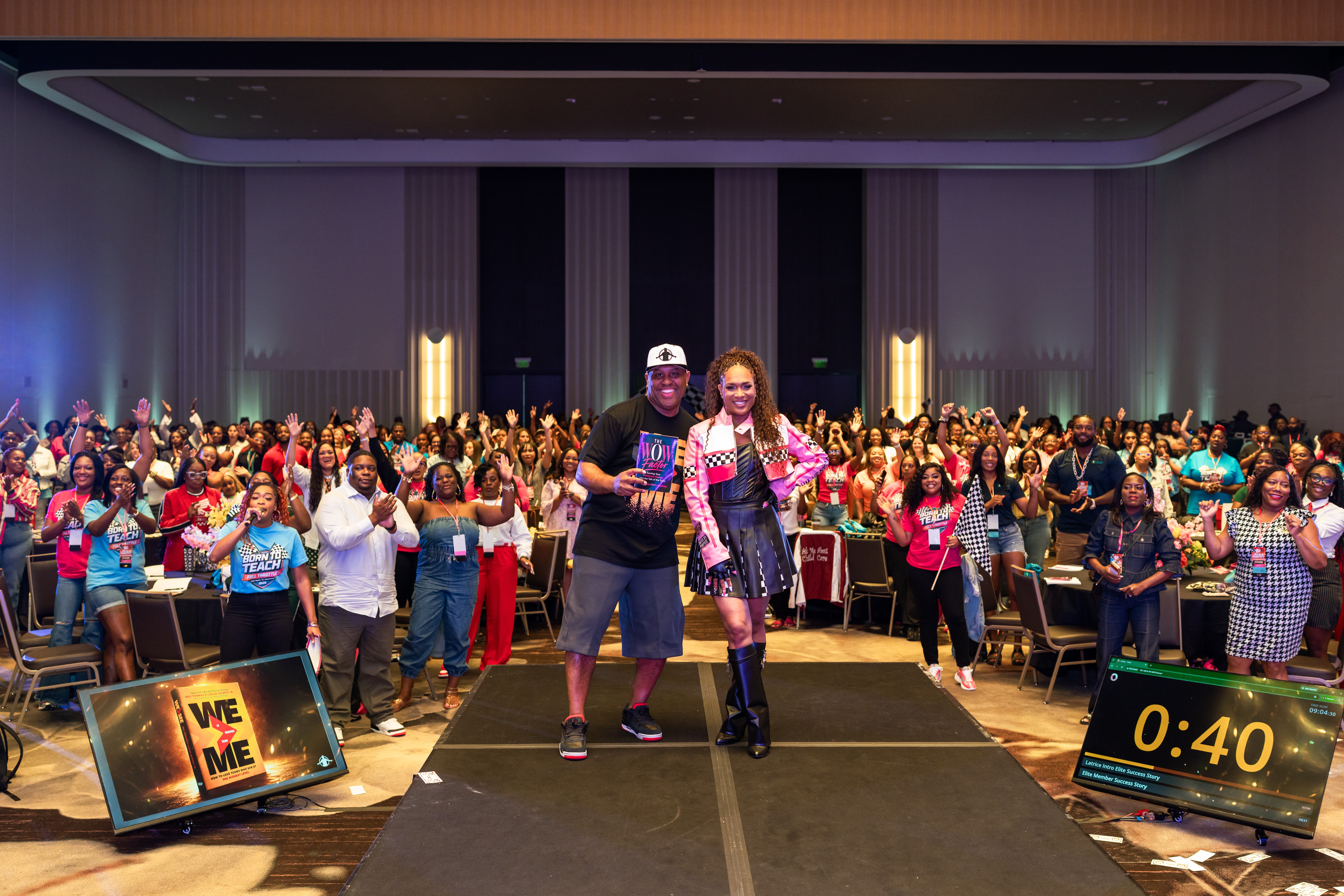 Two people walking on stage in front of a large audience, with a timer showing 40 seconds, and a sign reading 'WE ME'.