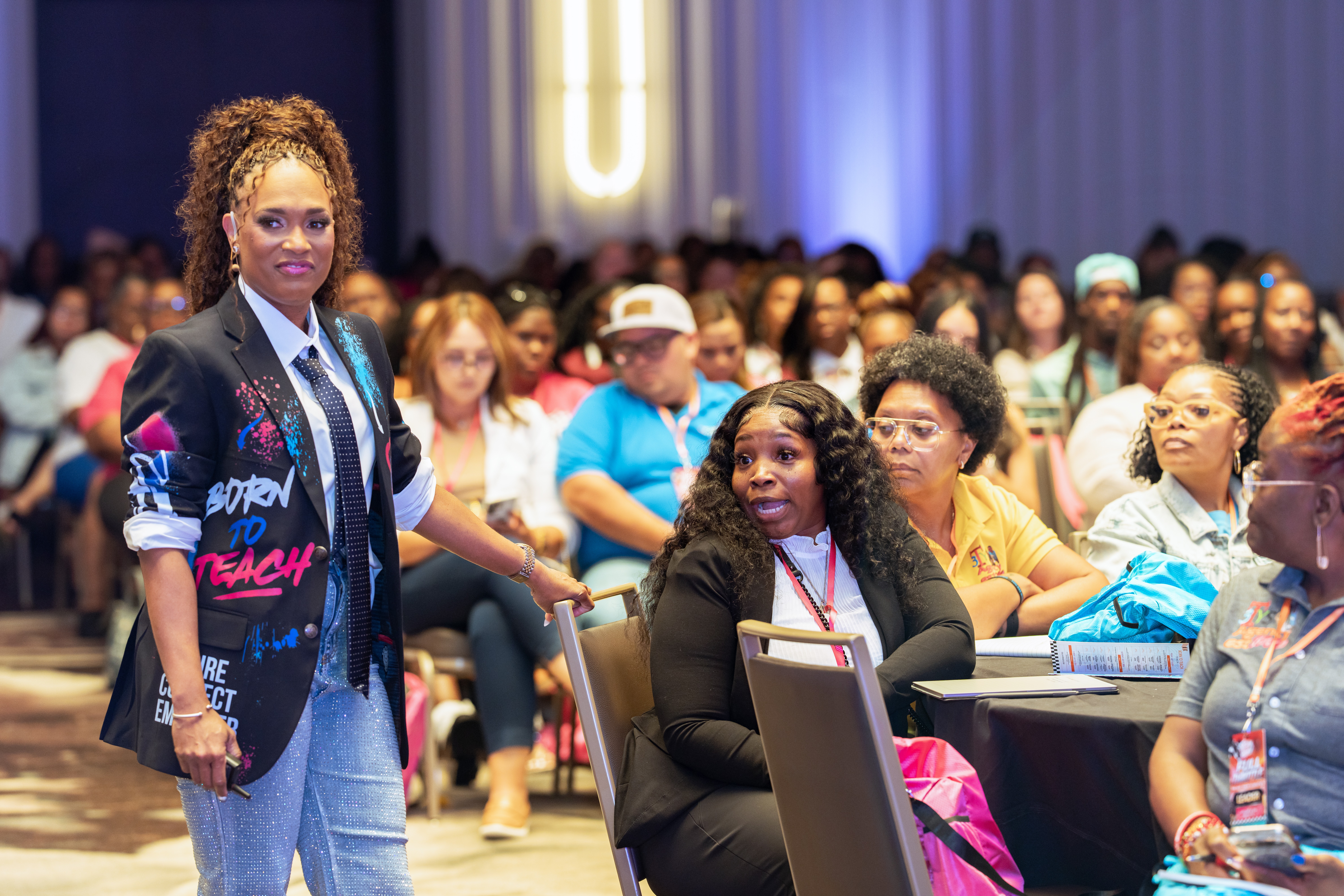 Woman standing in front of seated audience at conference or event, with diverse attendees in background.
