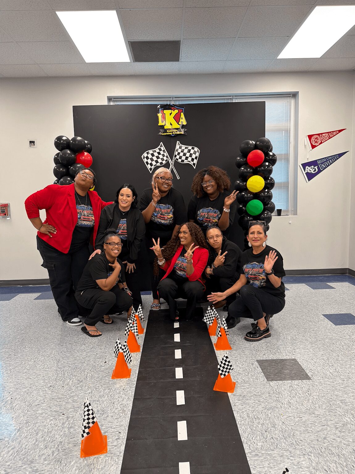 Group of eight women posing in front of a black backdrop with checkered flags, balloons, and a logo, inside a classroom.