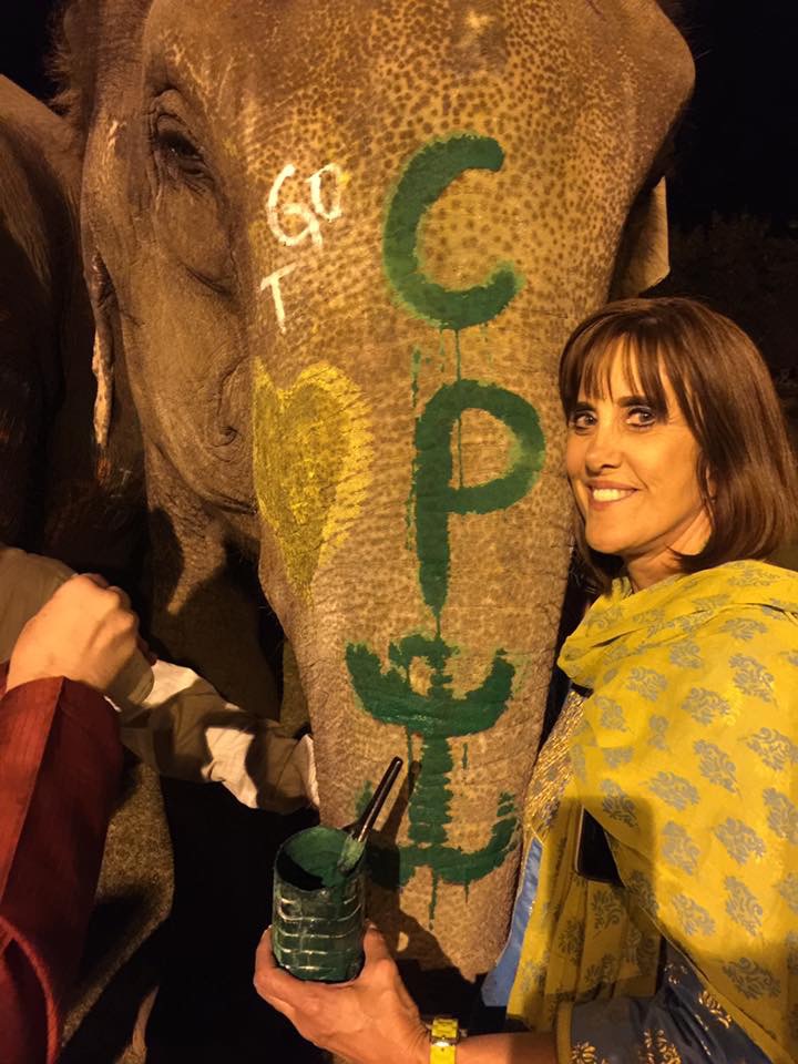 Woman smiling next to an elephant with painted letters and symbols on its face, holding a drink in a dark setting.