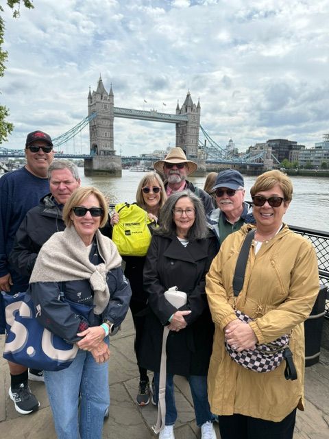 Group of people standing outdoors near a bridge, some wearing sunglasses and jackets, with cloudy sky in background.