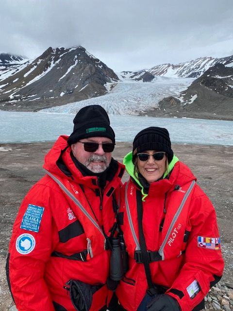 Two people in red jackets and black hats stand on snowy mountain terrain with snow-covered peaks in background.