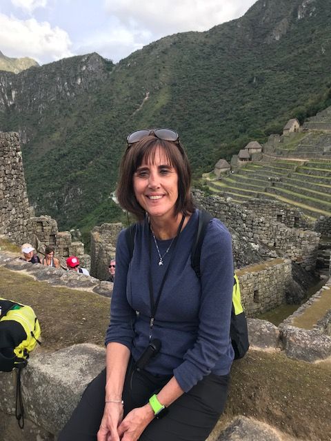 Woman with short hair, wearing a hat and navy jacket, sitting outdoors with ruins and green mountains in background.