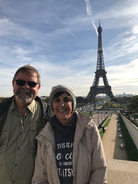Two people stand outdoors with the Eiffel Tower in the background, cloudy sky above, one wearing sunglasses and a jacket, the other with a cap and jacket.