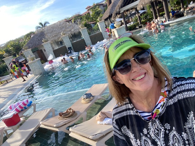 Smiling woman wearing sunglasses and a green cap at a poolside with people swimming and lounging in the background.