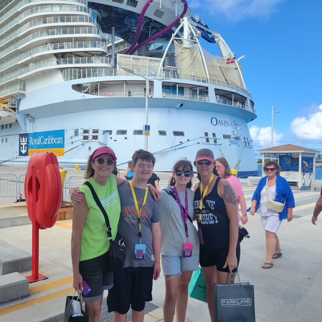 Group of five people standing in front of a large cruise ship, smiling, with a clear blue sky in the background.