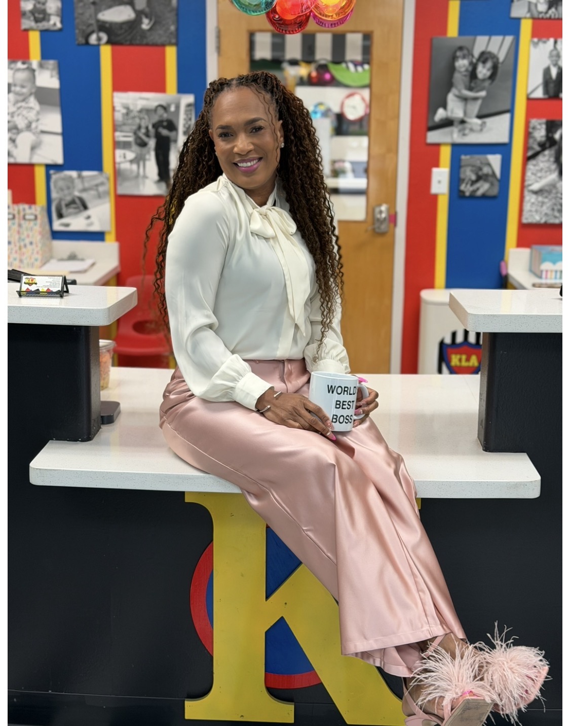 Woman sitting on a counter holding a mug, colorful background with photos and decorations, smiling, in a vibrant setting.
