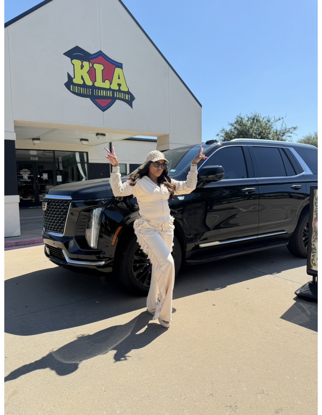 Woman in beige outfit and hat standing next to a black SUV outside a building with a KLA sign, waving with both hands.