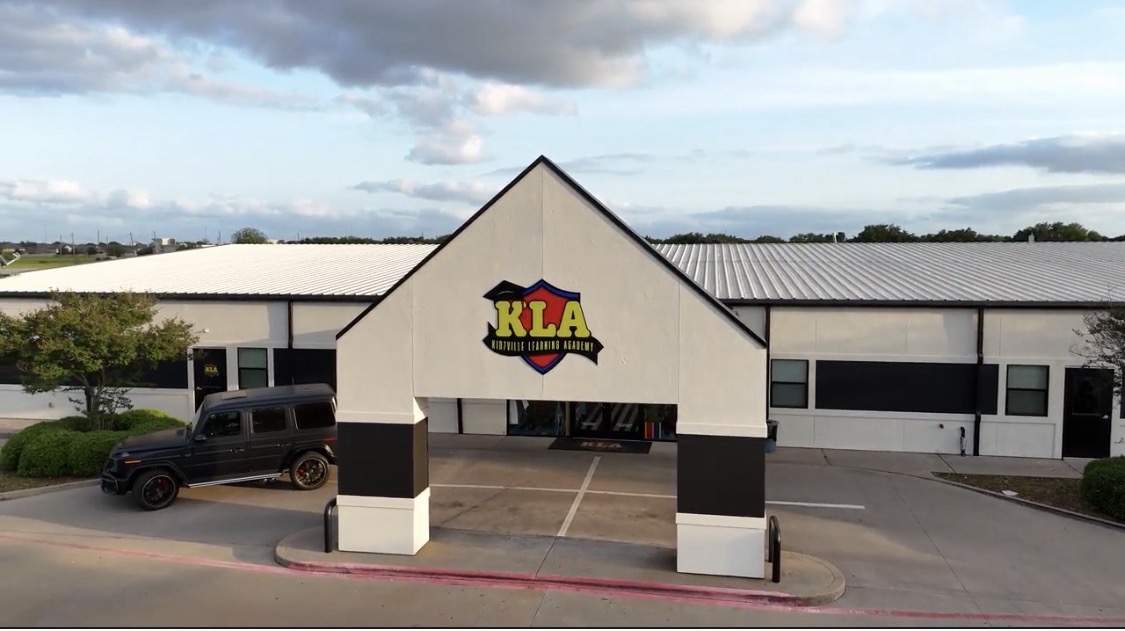Building with a sign reading 'KLA' and a parking lot in front, under a partly cloudy sky.