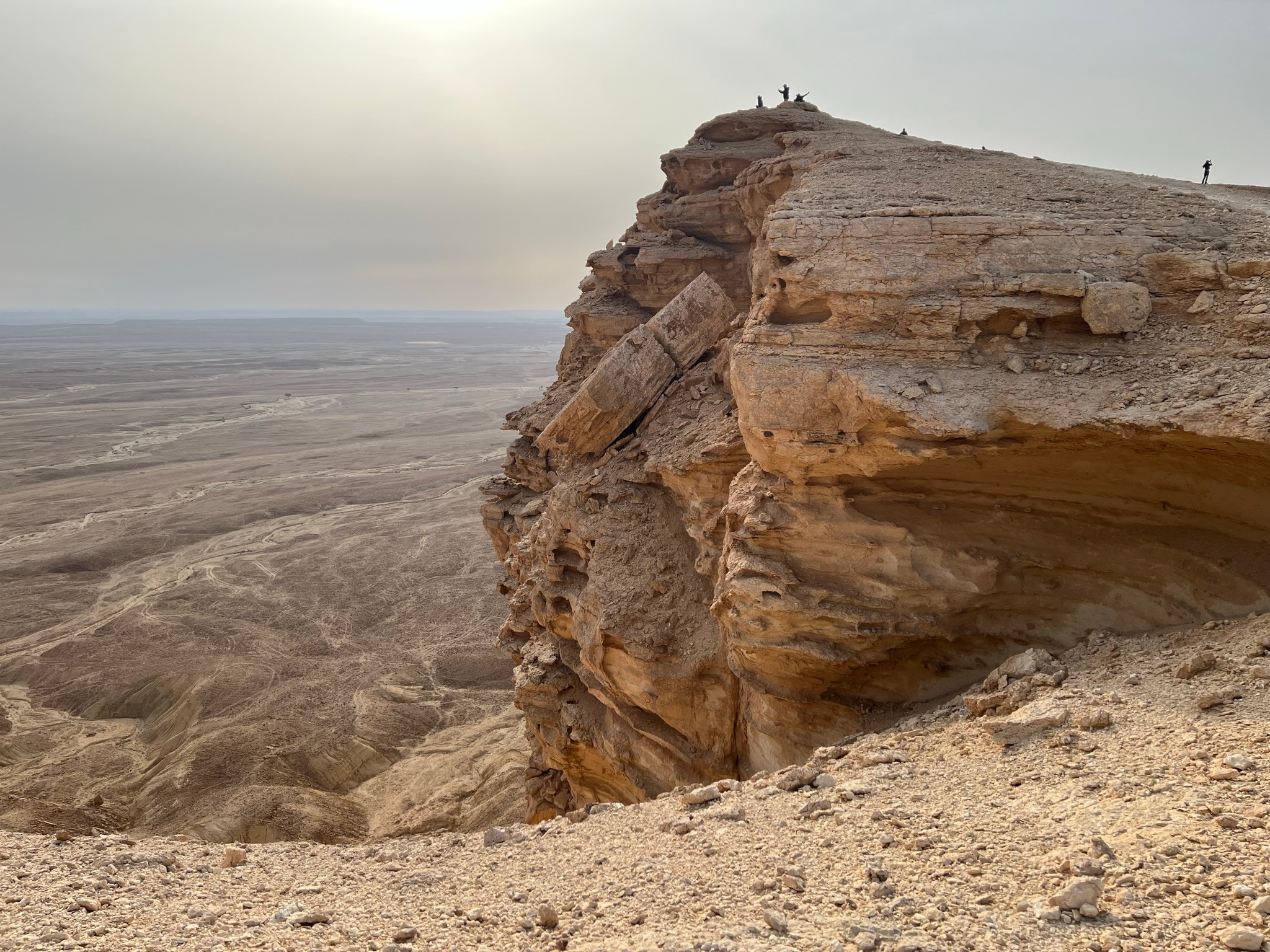Cliff with layered rock formation overlooking a desert landscape, with people standing at the top, under a bright sky.
