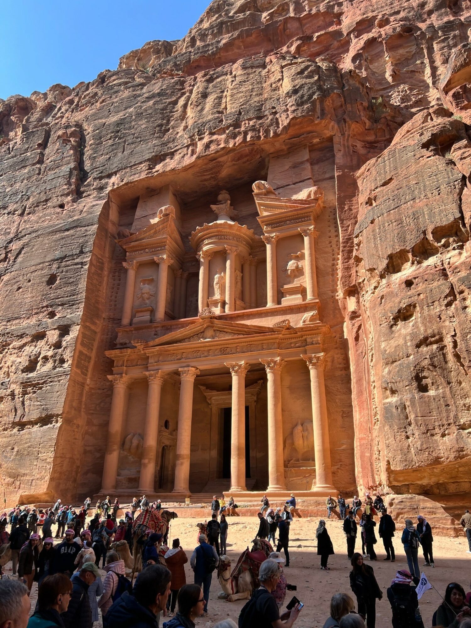 Ancient rock-cut structure with multiple columns and detailed carvings, set against a cliff face, with a large crowd of visitors below.
