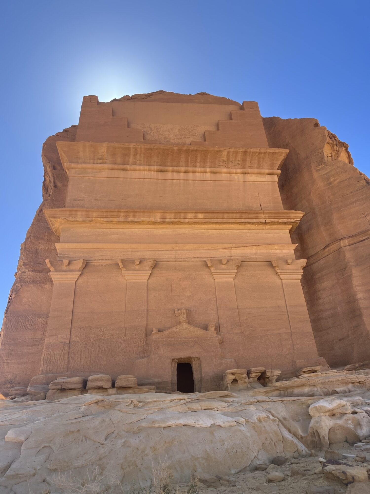 Ancient stone structure with a small entrance, set against a clear blue sky, sunlight illuminating the top, rocky ground in foreground.